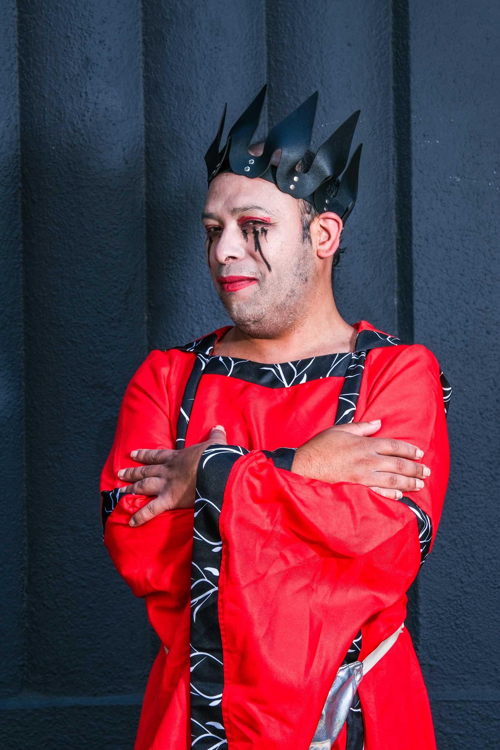 A person in red and black attire, wearing a spiked crown, with theatrical makeup featuring exaggerated red eyeshadow and black tears, stands against a dark wall.