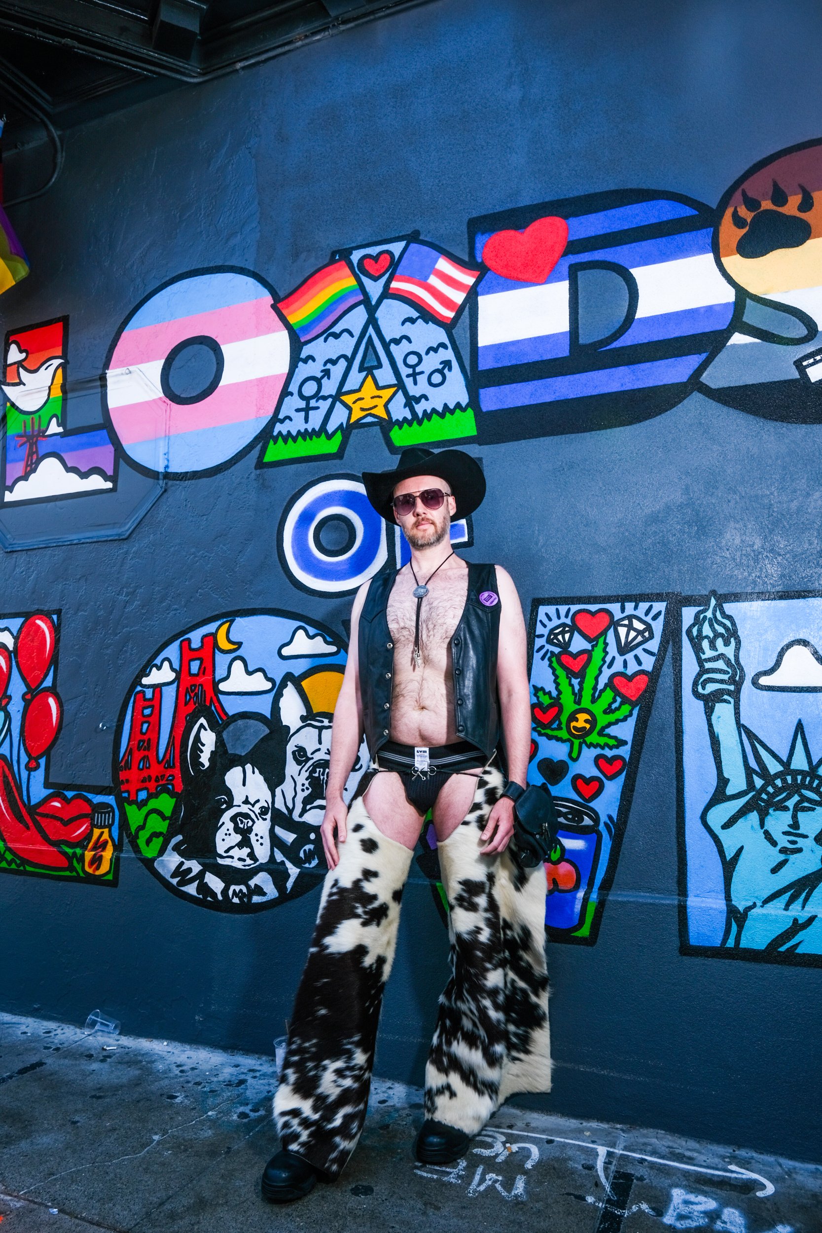 A person in a cowboy hat, sunglasses, and leather vest stands in front of a colorful mural with the word &quot;LOVE&quot; incorporating various pride symbols.