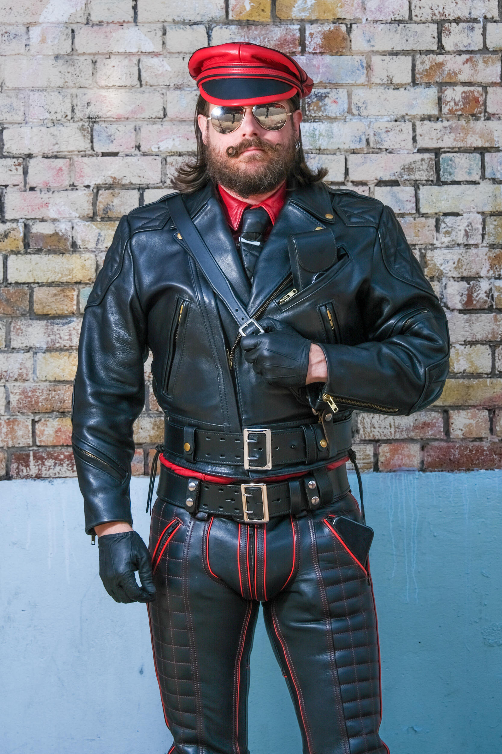 A man with a mustache and sunglasses wears a black leather jacket, pants with red trim, gloves, a red-topped cap, and multiple belts, standing against a brick wall.