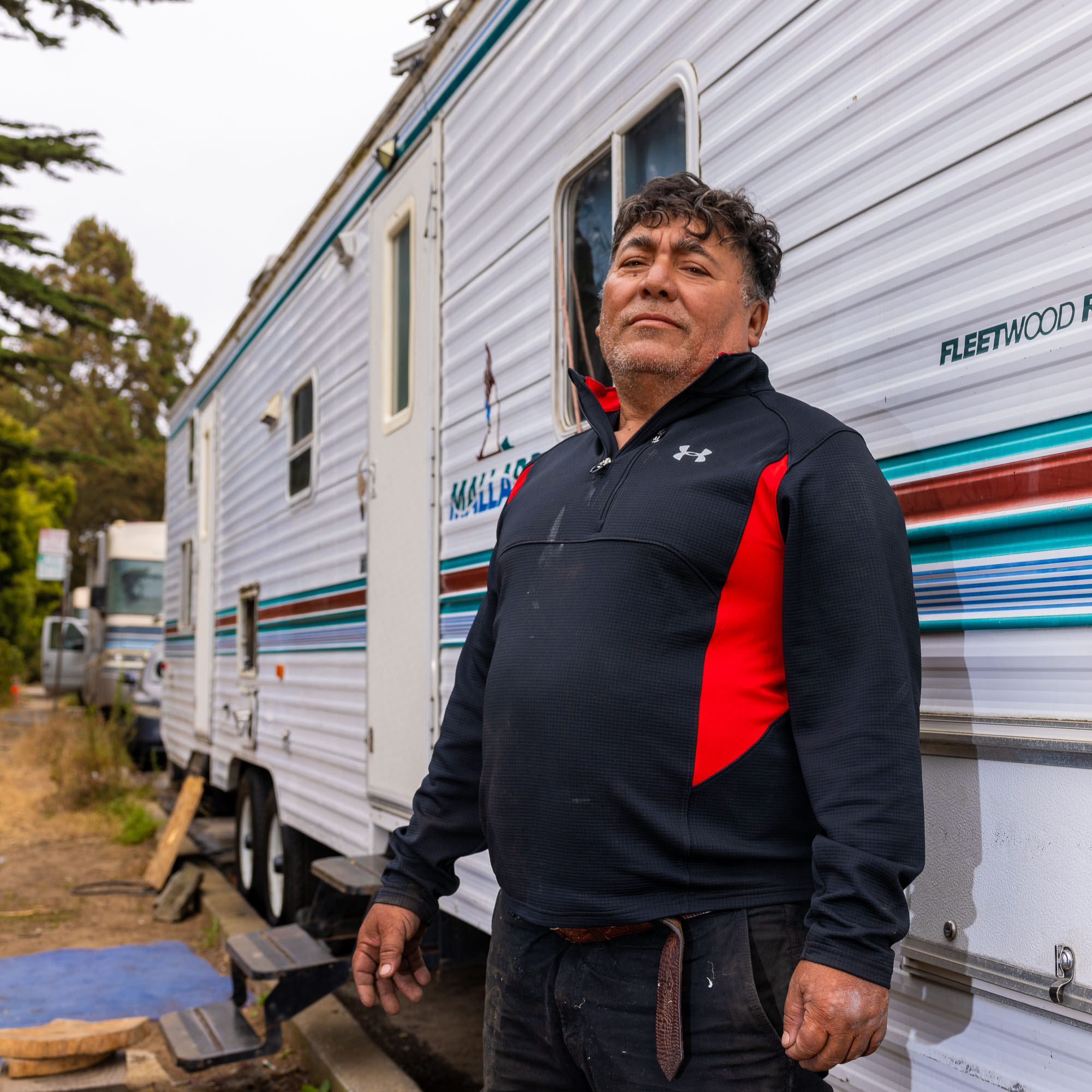 A man in a black and red jacket stands confidently in front of a white RV with striped detailing. Trees and other RVs can be seen in the background.