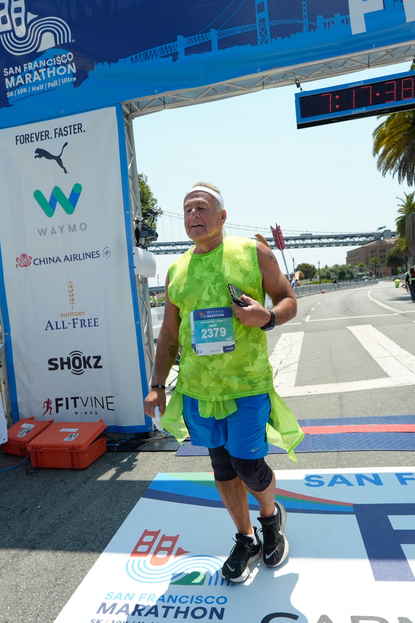 A runner wearing a green shirt crosses the finish line at the San Francisco Marathon under a blue arch with sponsor logos and a timing clock overhead.