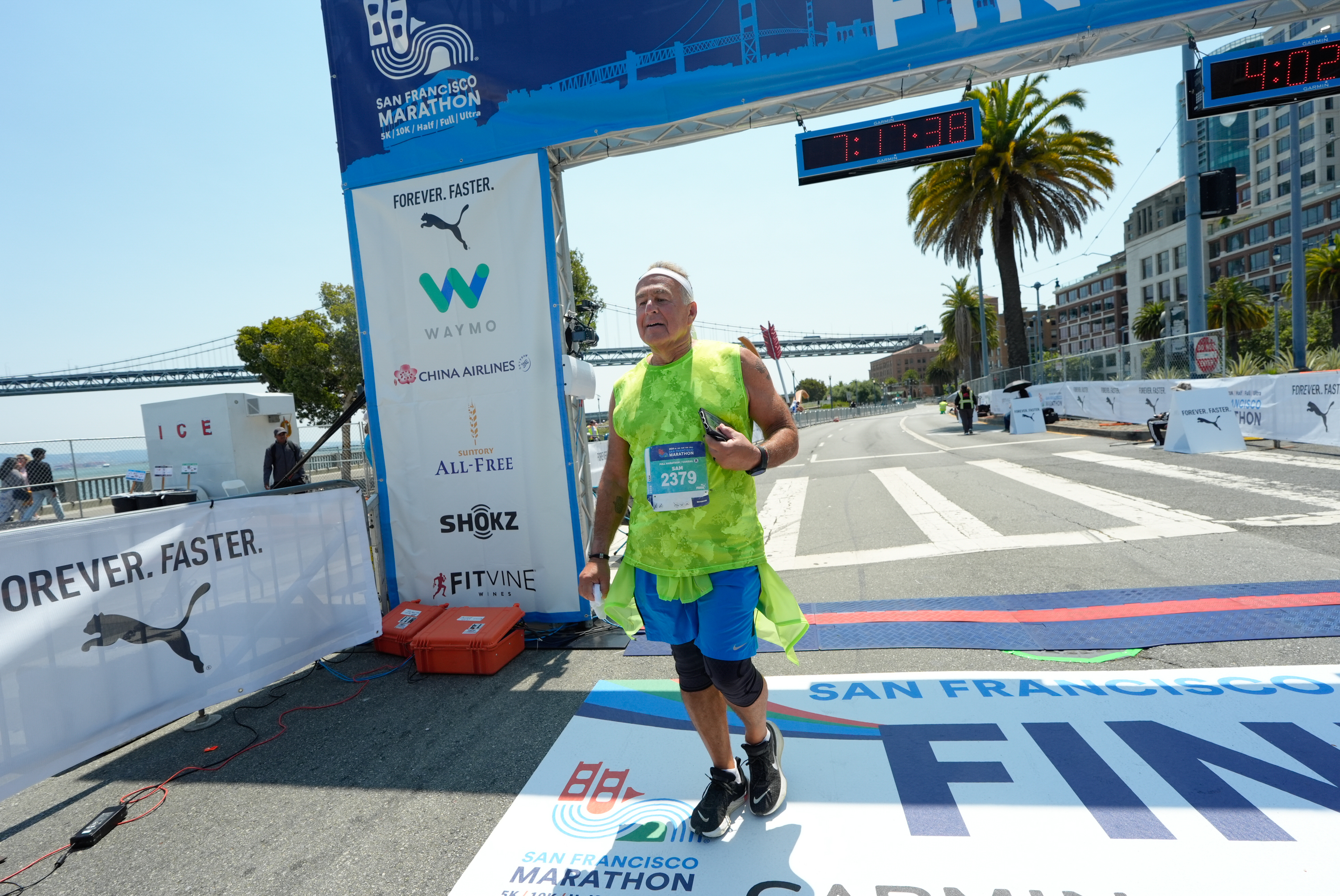 A runner wearing a green shirt crosses the finish line at the San Francisco Marathon under a blue arch with sponsor logos and a timing clock overhead.