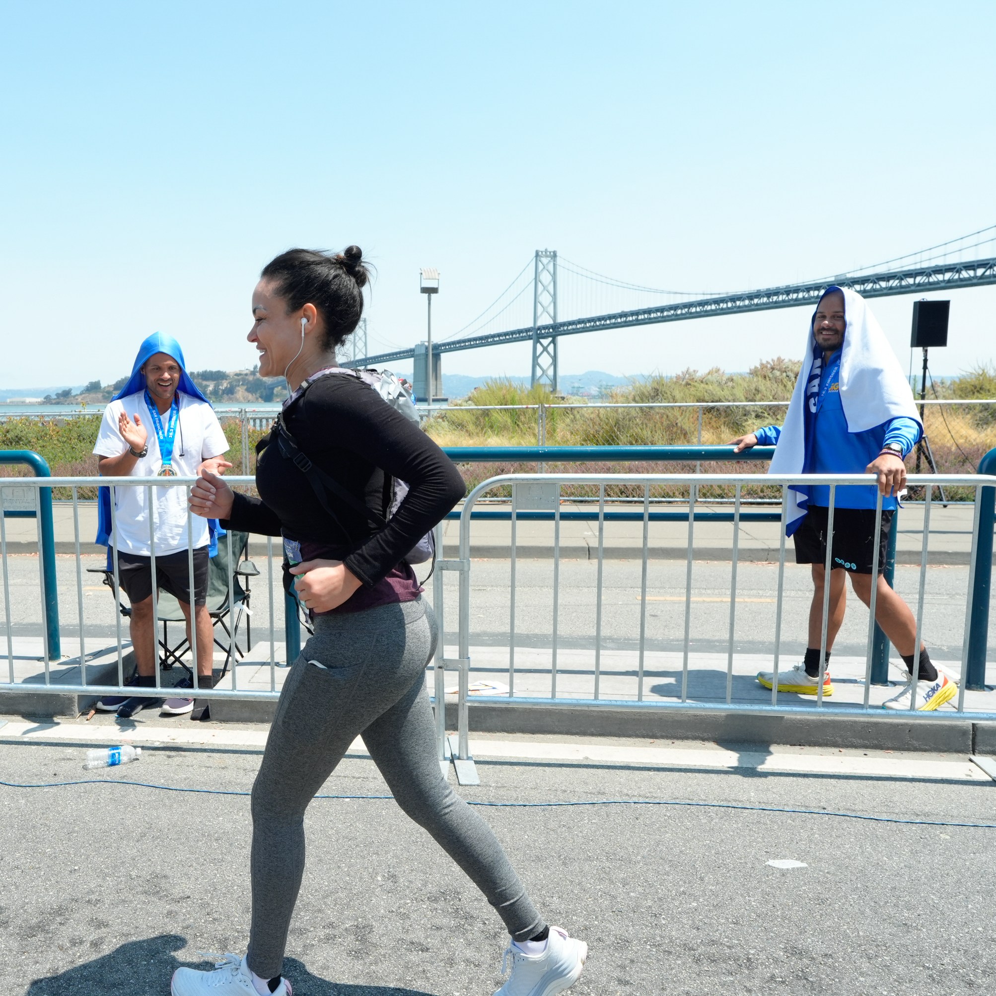 A woman jogs in a race with earbuds on, while two men with towels over their heads cheer her on behind a barrier near a bridge on a sunny day.