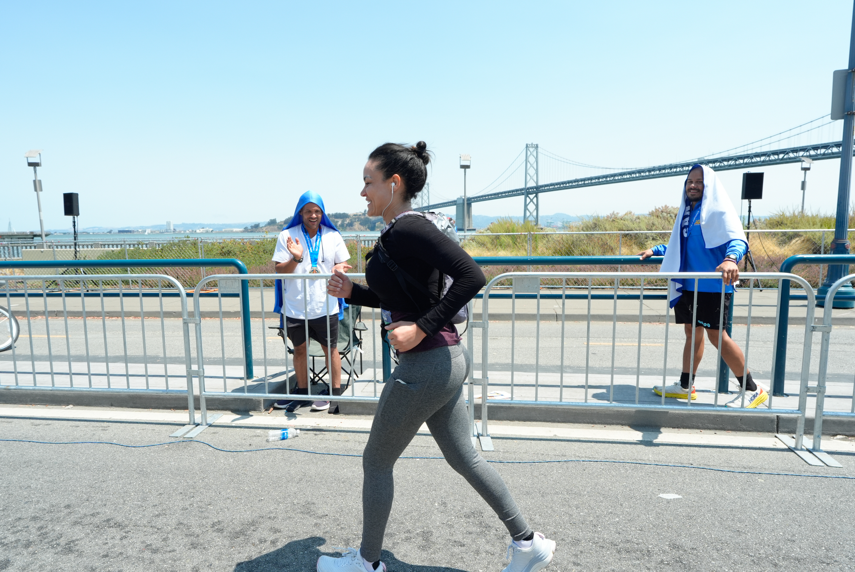 A woman jogs in a race with earbuds on, while two men with towels over their heads cheer her on behind a barrier near a bridge on a sunny day.