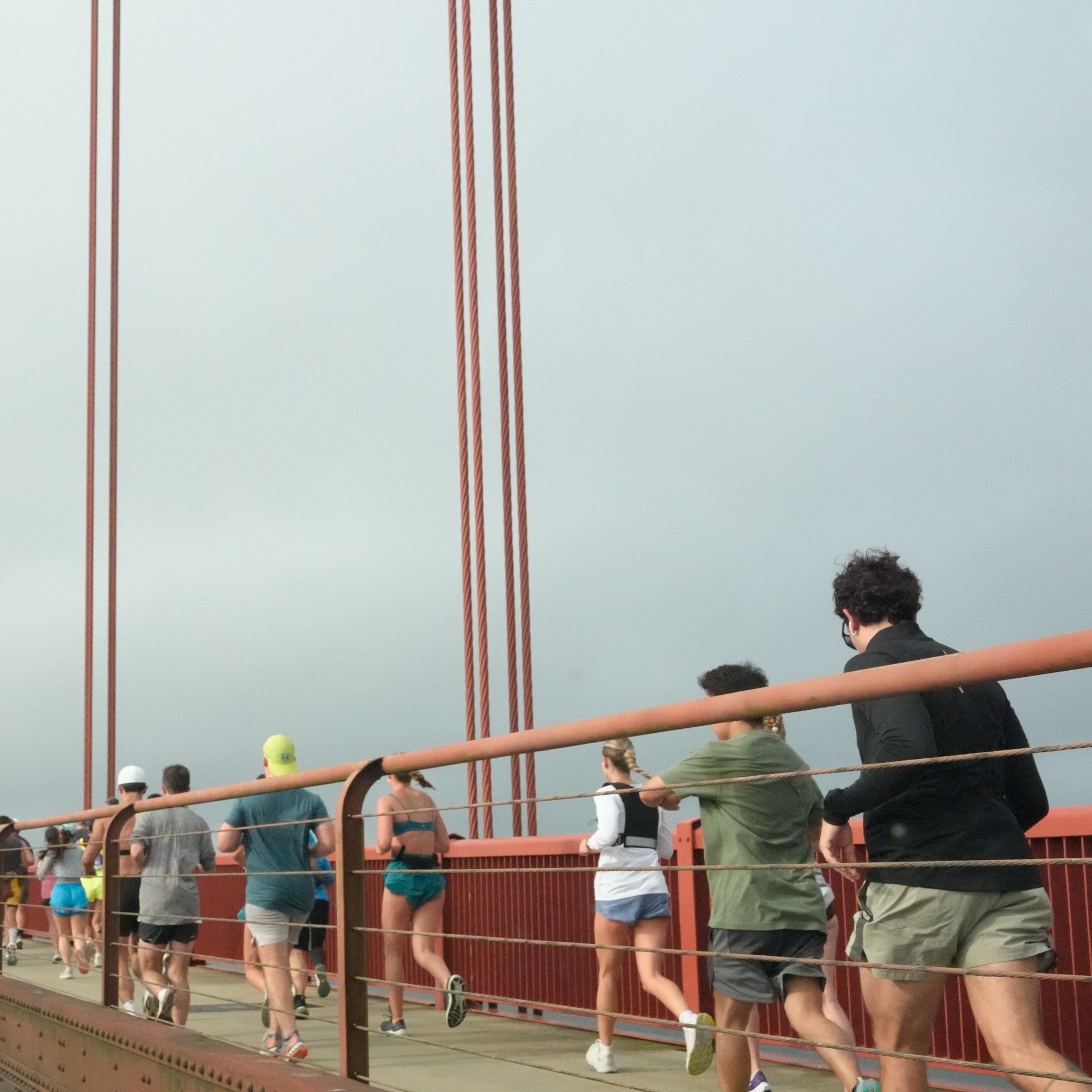 People are jogging along the pedestrian path of a large red suspension bridge on a cloudy day, while cars drive on the adjacent road.