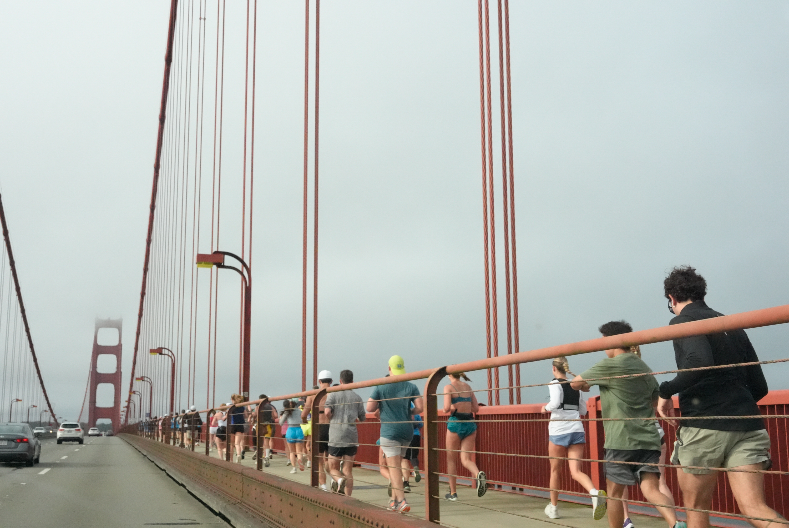 People are jogging along the pedestrian path of a large red suspension bridge on a cloudy day, while cars drive on the adjacent road.