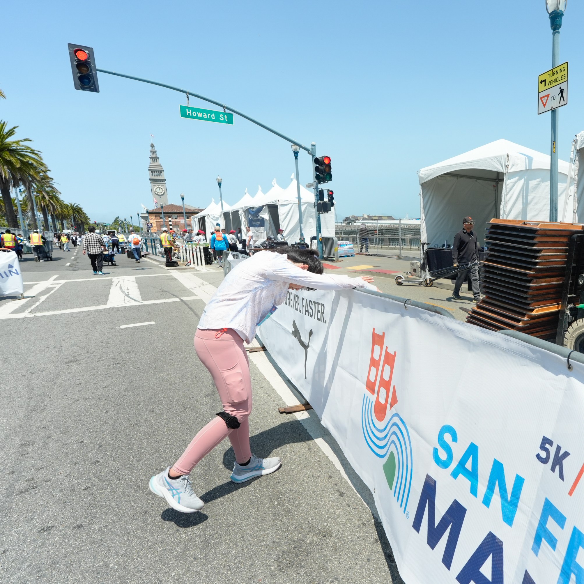 A person in pink pants leans on a barrier while a worker on a forklift carries tables. The scene is near a &quot;San Francisco Marathon&quot; sign on Howard Street.