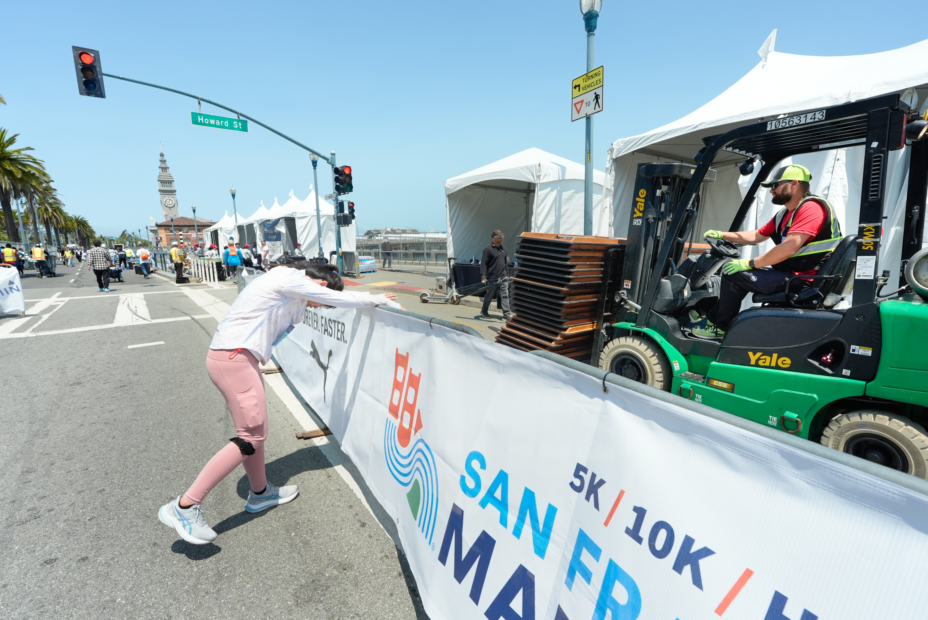 A person in pink pants leans on a barrier while a worker on a forklift carries tables. The scene is near a &quot;San Francisco Marathon&quot; sign on Howard Street.