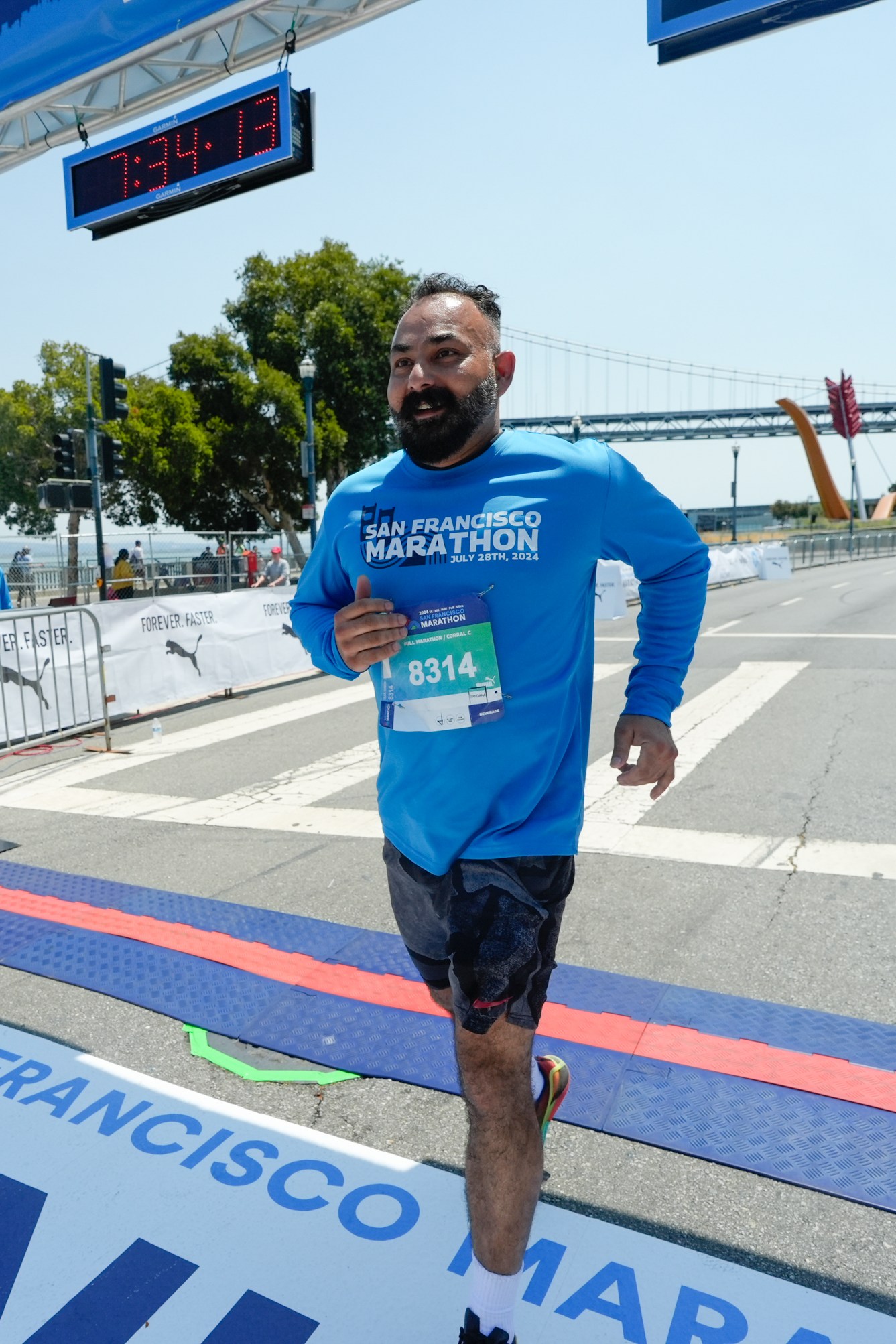 A man in a blue &quot;San Francisco Marathon&quot; shirt crosses the marathon finish line, with a digital clock above and a bridge in the background.