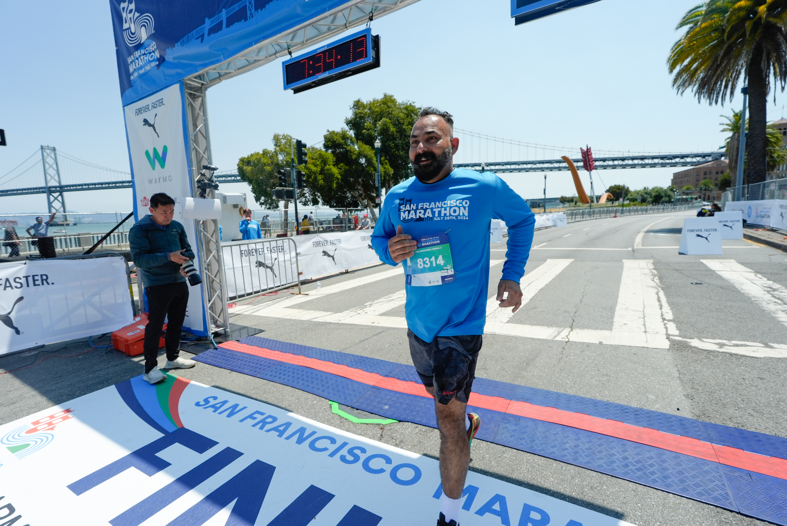 A man in a blue &quot;San Francisco Marathon&quot; shirt crosses the marathon finish line, with a digital clock above and a bridge in the background.