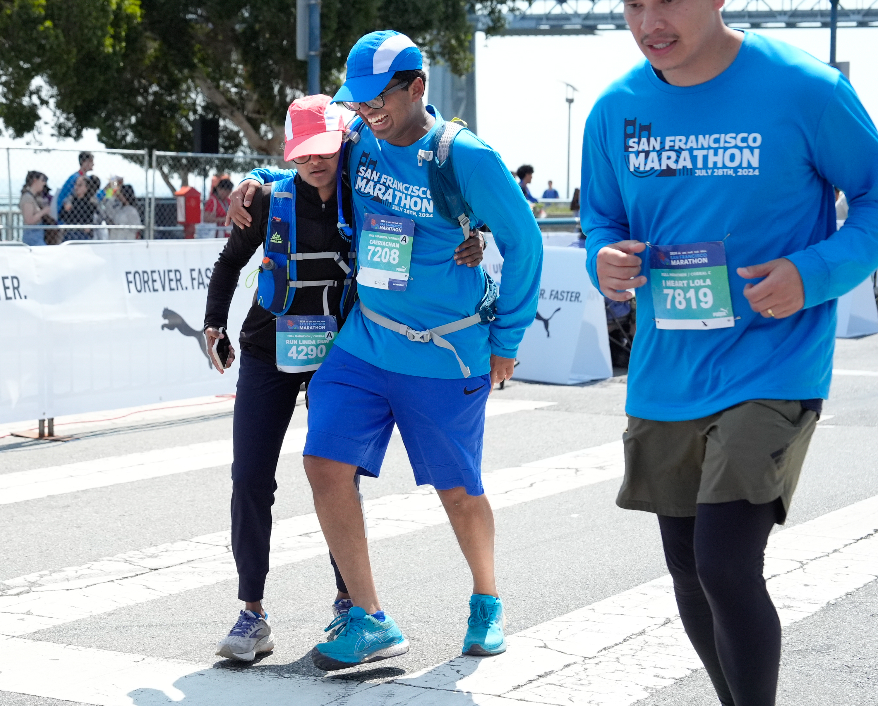 Three runners wearing blue “San Francisco Marathon” shirts cross the finish line together, two of them assisting each other while smiling.