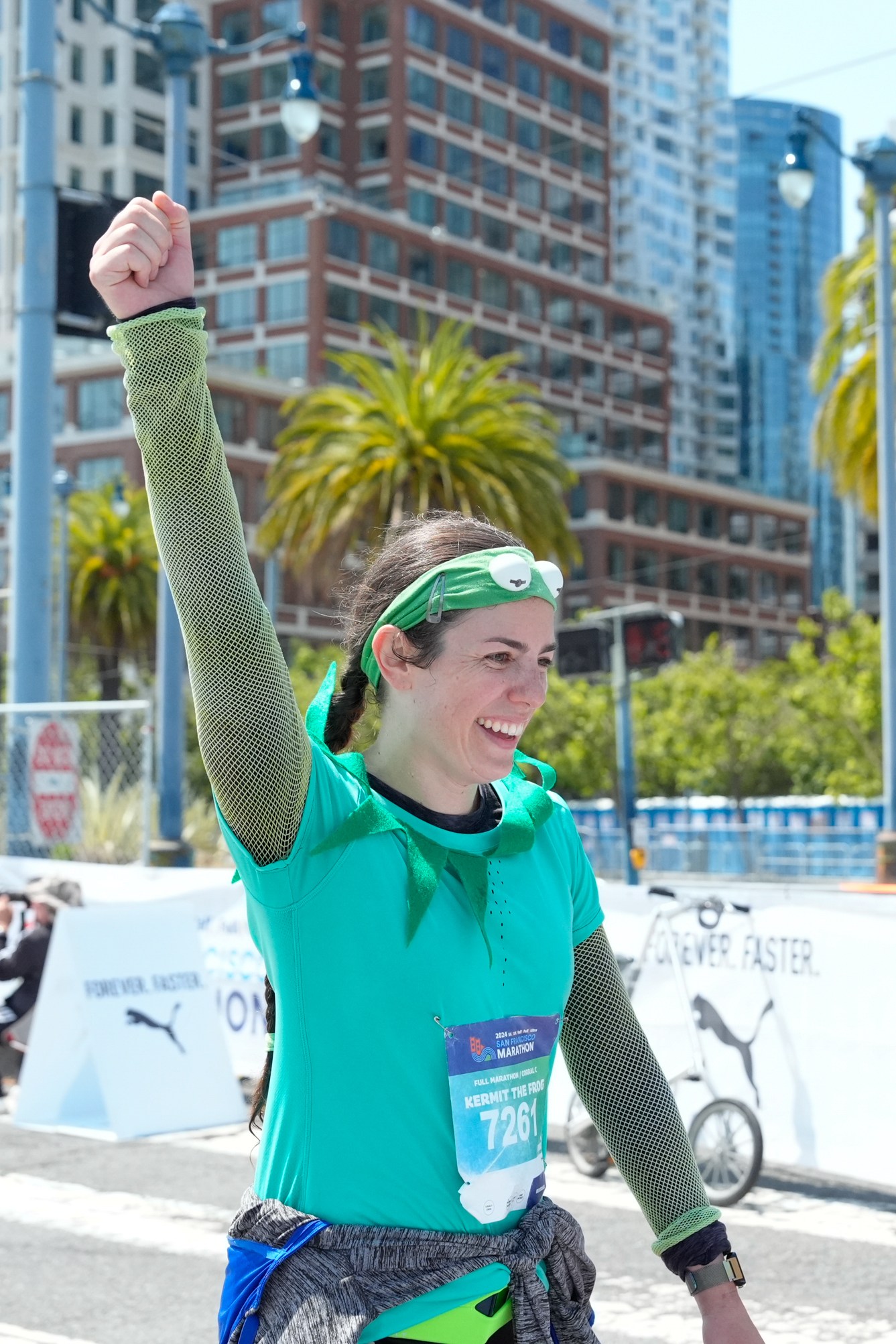 A woman in a teal outfit raises her fist in victory, smiling, with a race bib number &quot;7261.&quot; She's on a city street, with palm trees and tall buildings in the background.