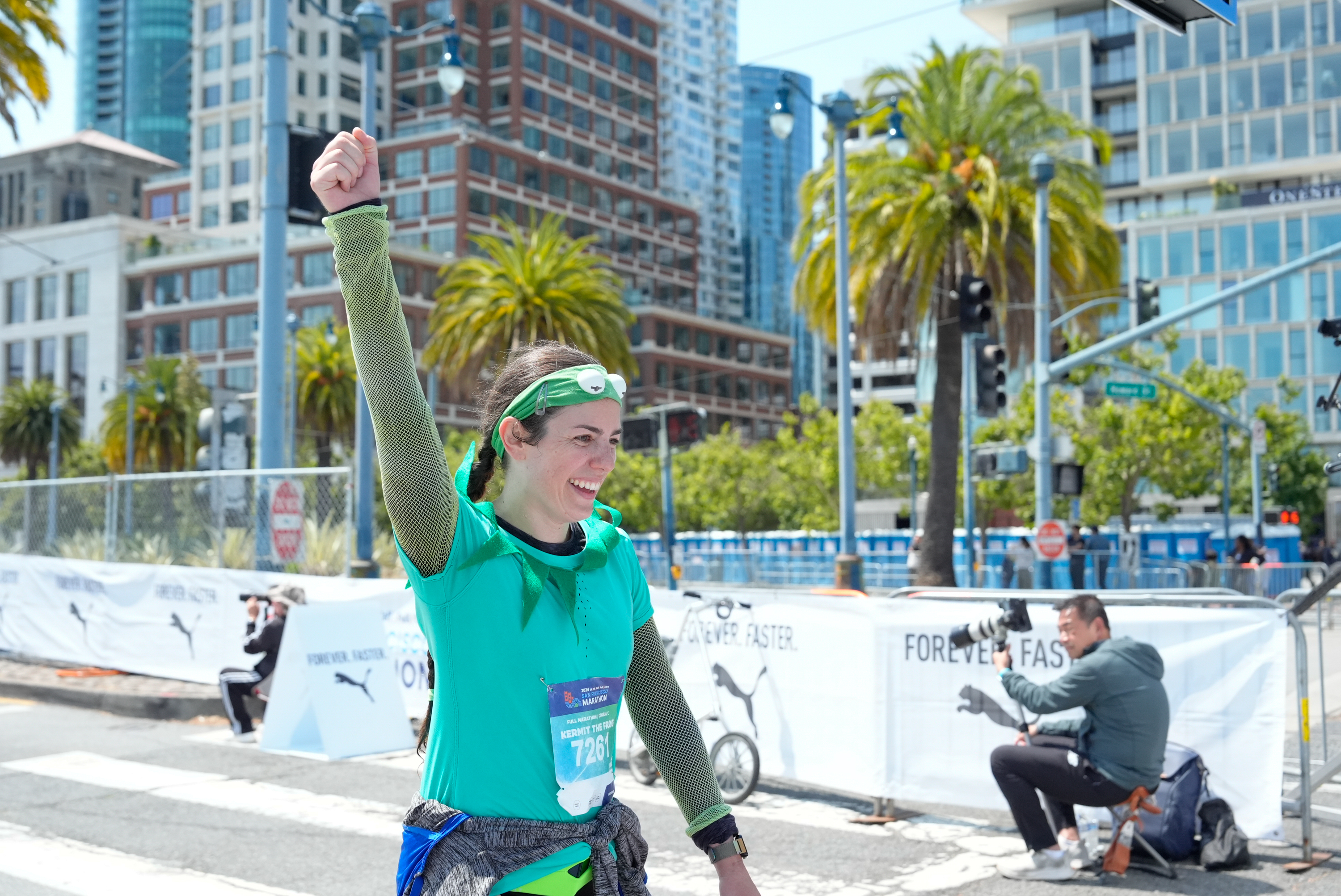 A woman in a teal outfit raises her fist in victory, smiling, with a race bib number &quot;7261.&quot; She's on a city street, with palm trees and tall buildings in the background.