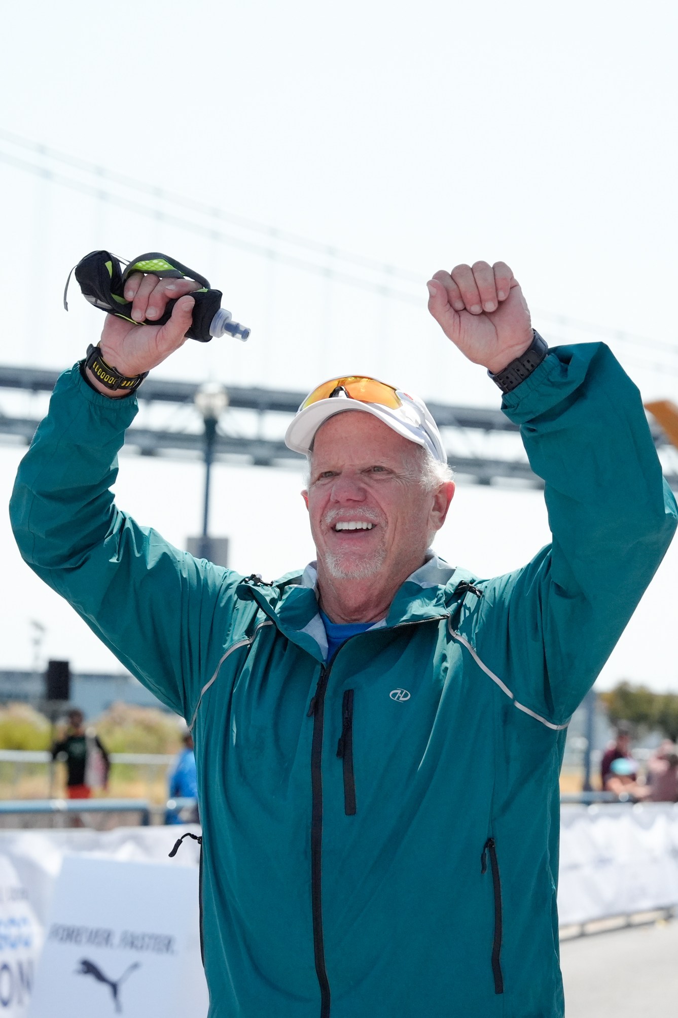 A man in a teal jacket and white cap raises his arms in victory at a marathon finish line, with a bridge and arrow sculpture in the background.