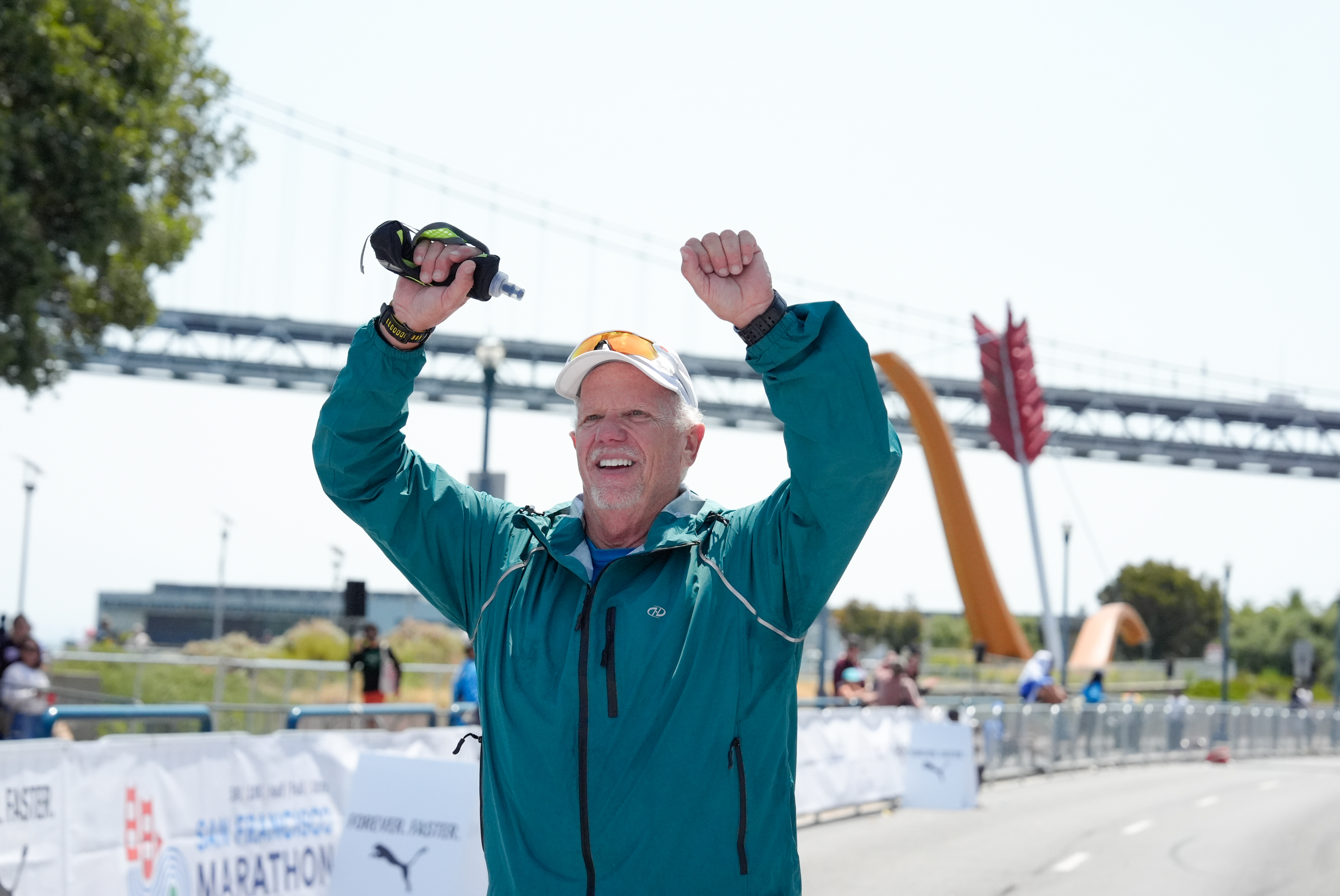 A man in a teal jacket and white cap raises his arms in victory at a marathon finish line, with a bridge and arrow sculpture in the background.