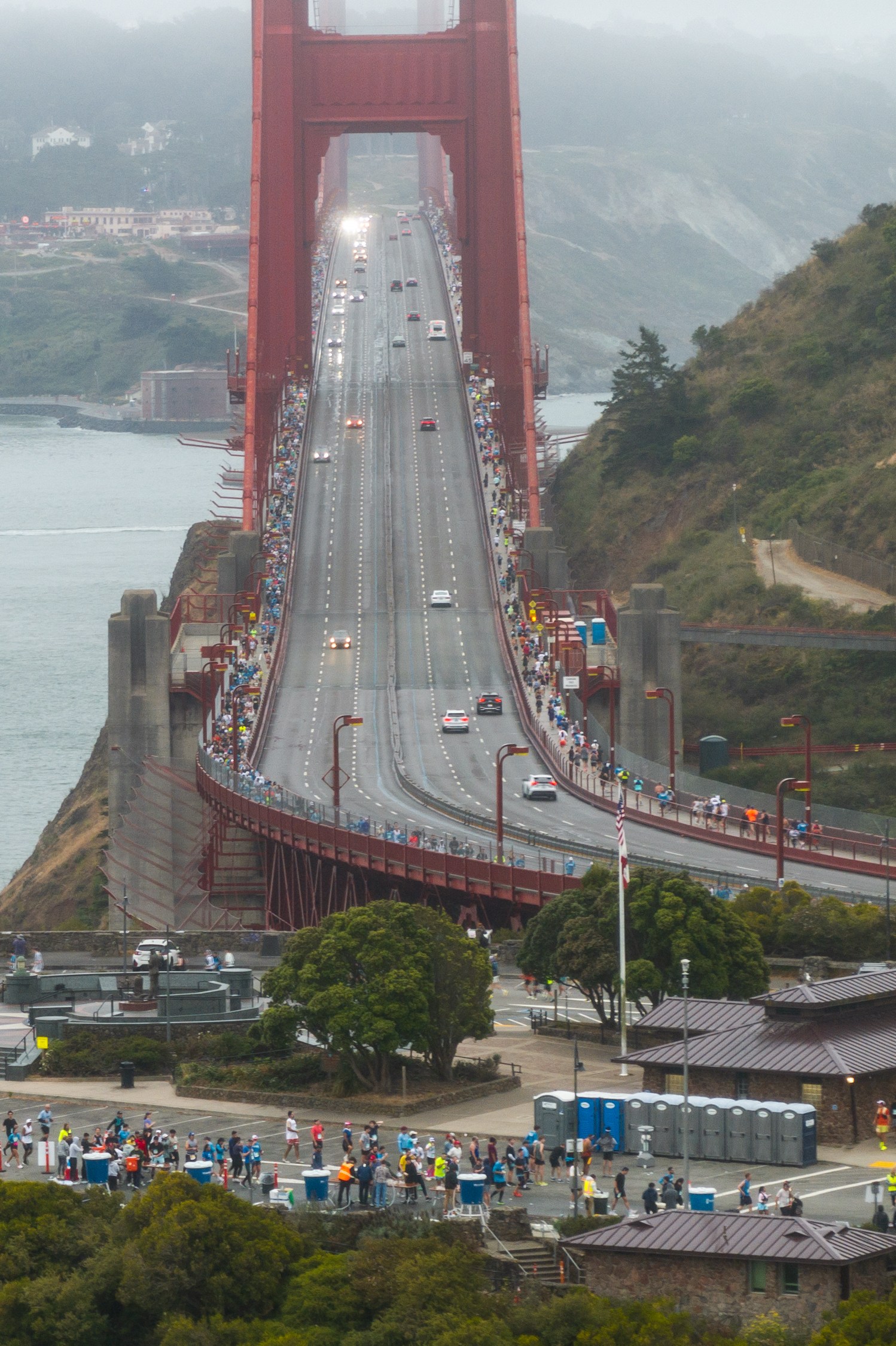 A large group of people is gathered at one end of the Golden Gate Bridge, with some cars driving across it. The scene overlooks water and a foggy, tree-filled background.