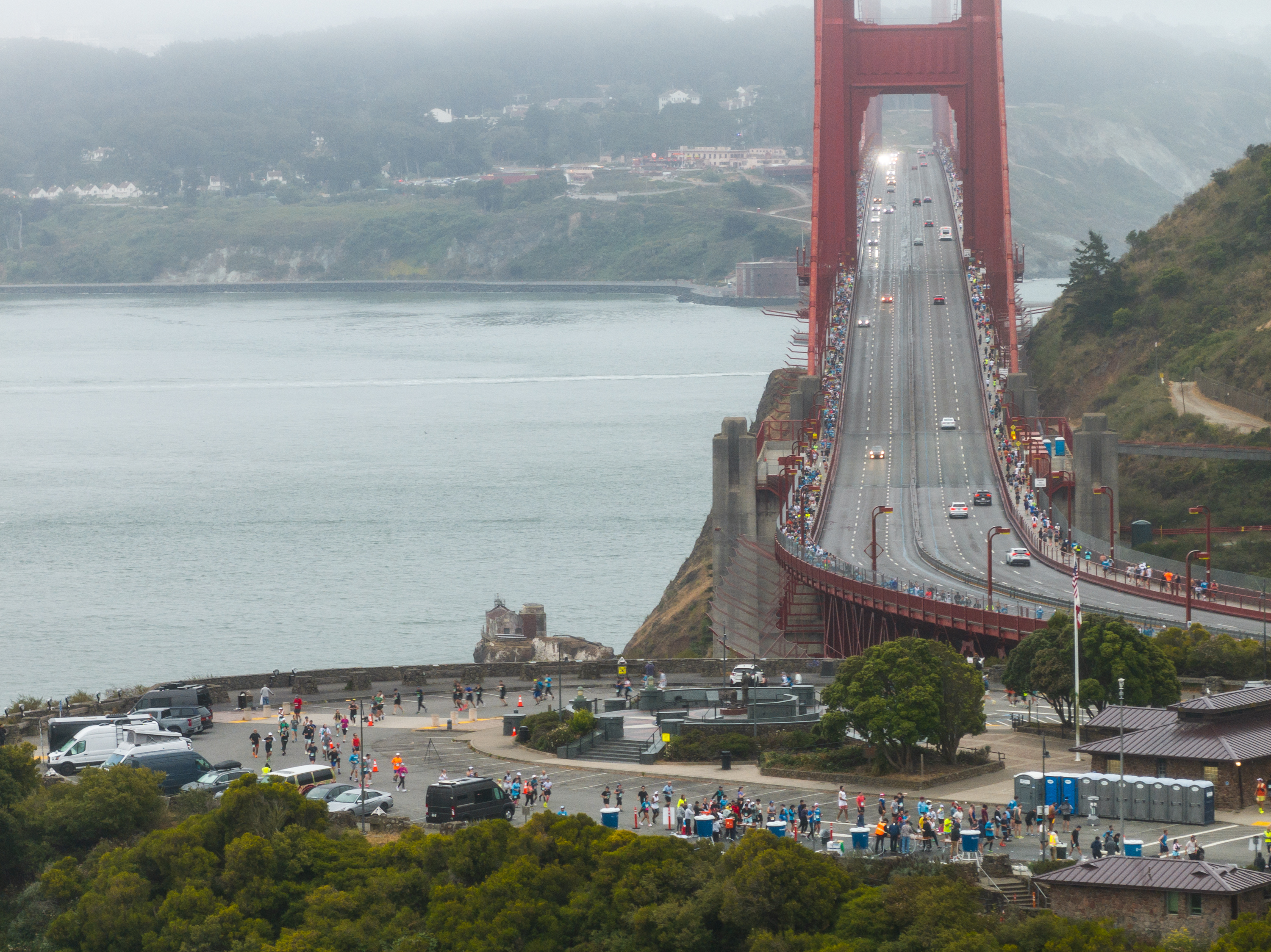 A large group of people is gathered at one end of the Golden Gate Bridge, with some cars driving across it. The scene overlooks water and a foggy, tree-filled background.