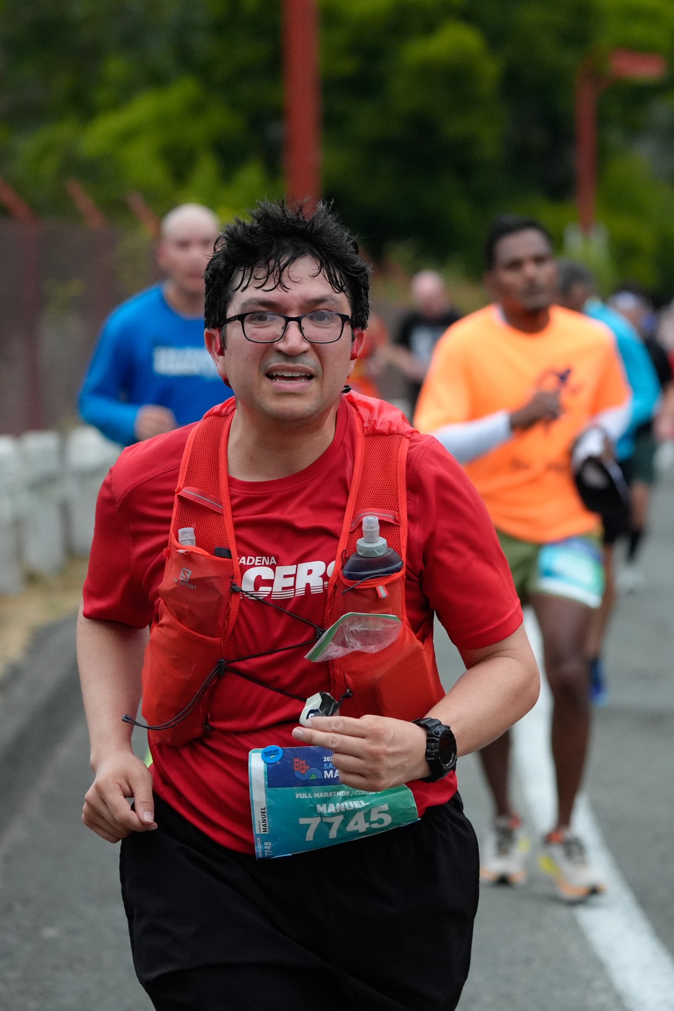 A man wearing a red shirt, glasses, and a water bottle vest runs in a marathon with other runners in the background on a road lined with traffic cones. 