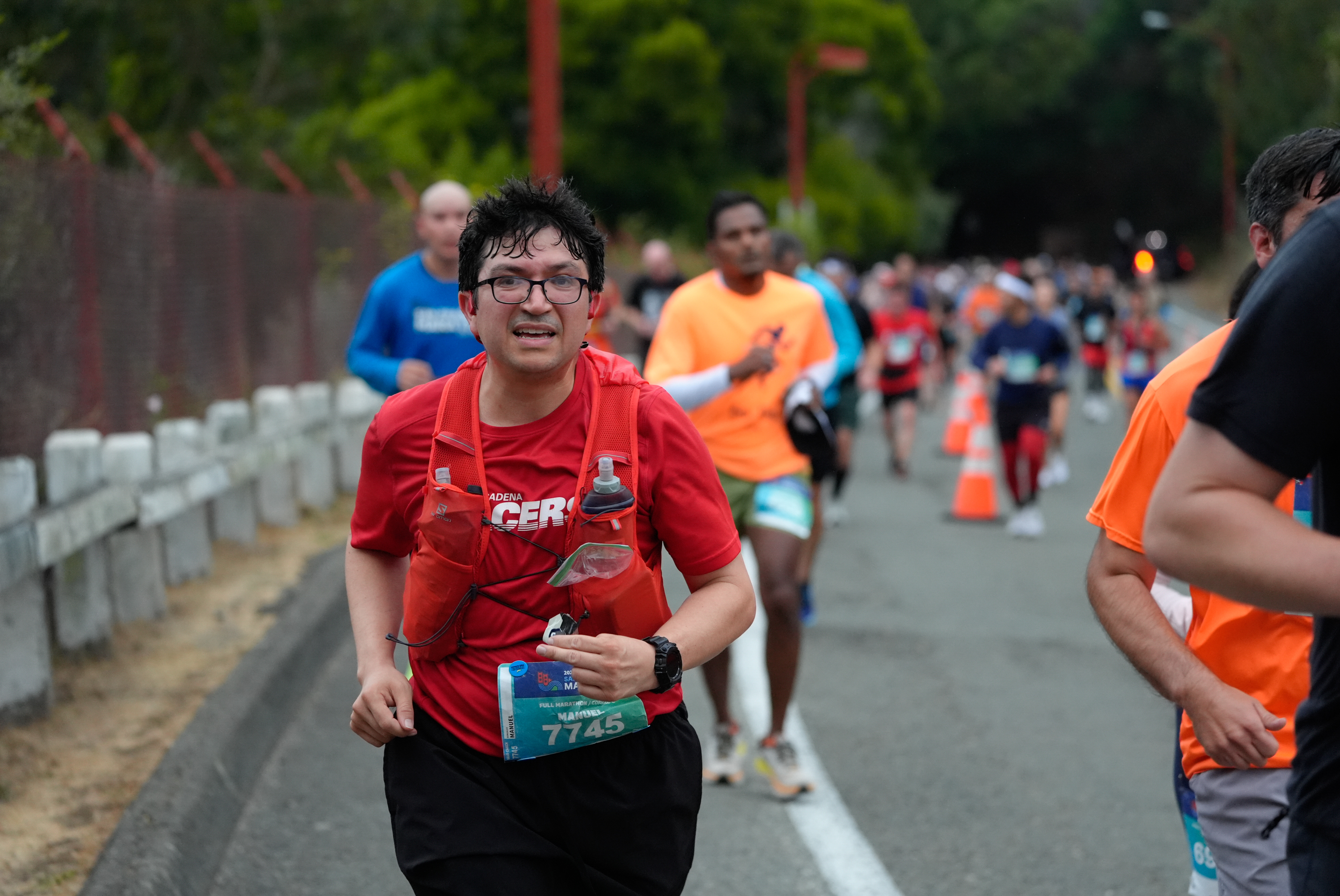 A man wearing a red shirt, glasses, and a water bottle vest runs in a marathon with other runners in the background on a road lined with traffic cones. 