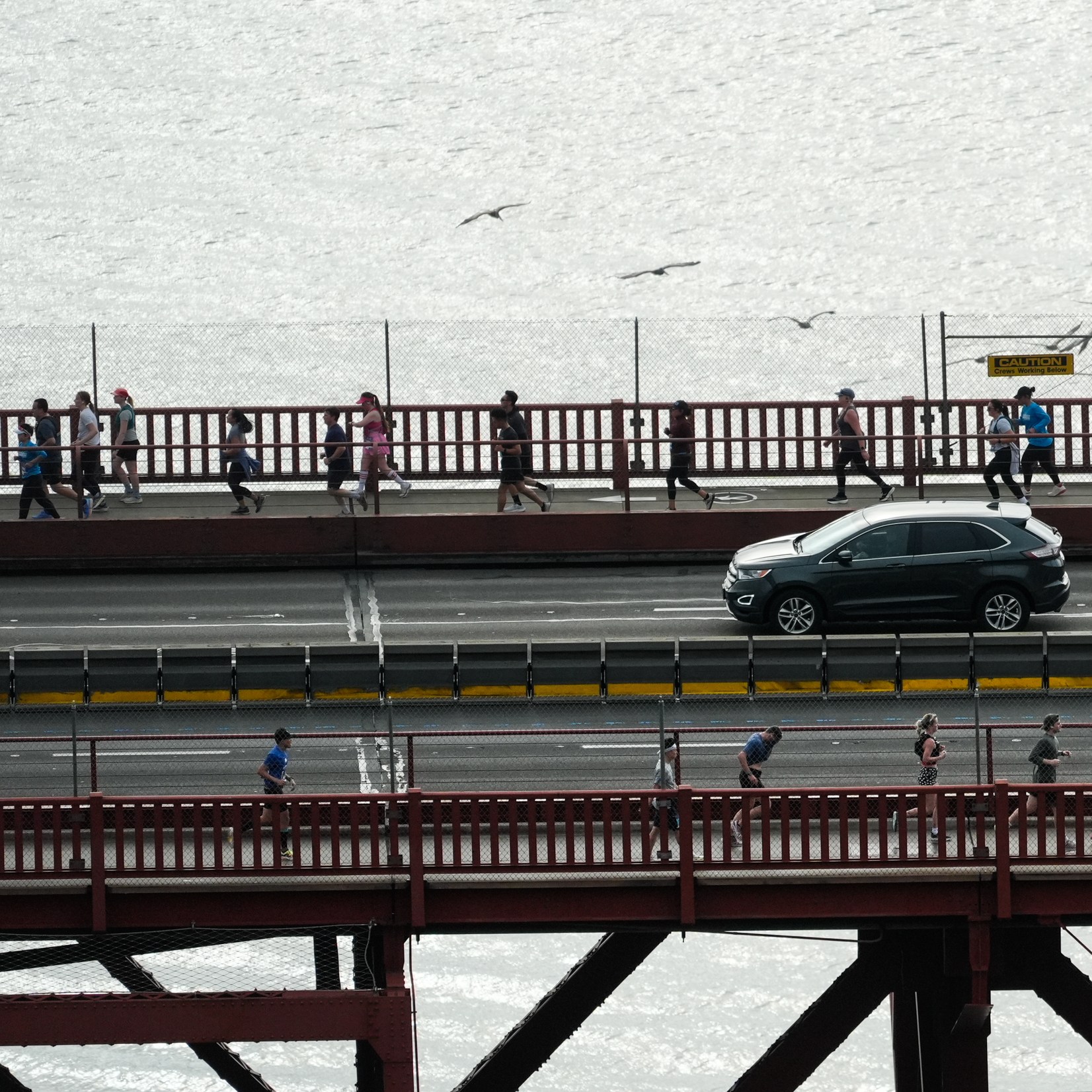 People are walking on two levels of a red bridge, separated by a road with a black SUV. Seagulls are flying in the background.