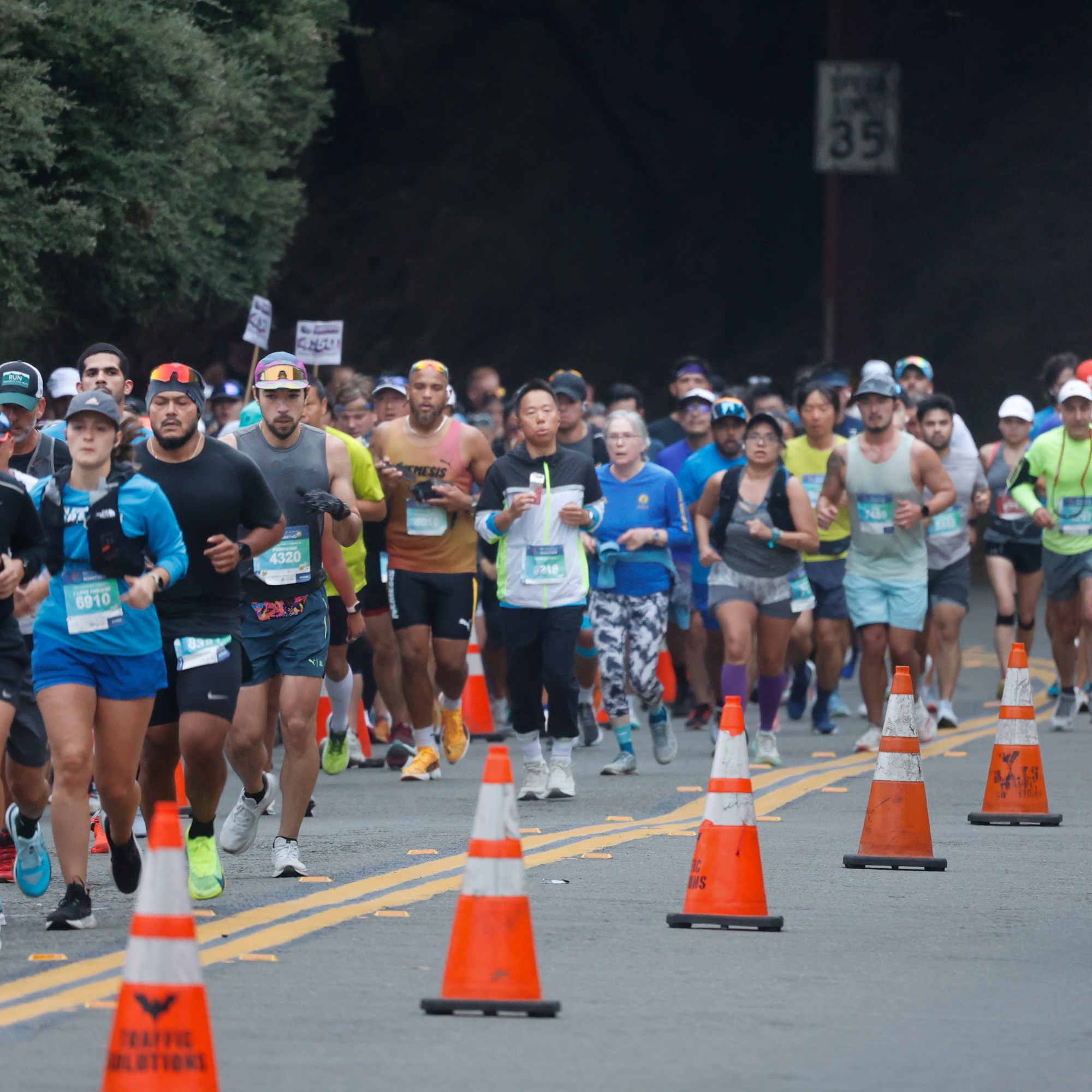 A group of runners in athletic gear is participating in a race on a road with traffic cones, while a motorbike rider and car are visible to the right.