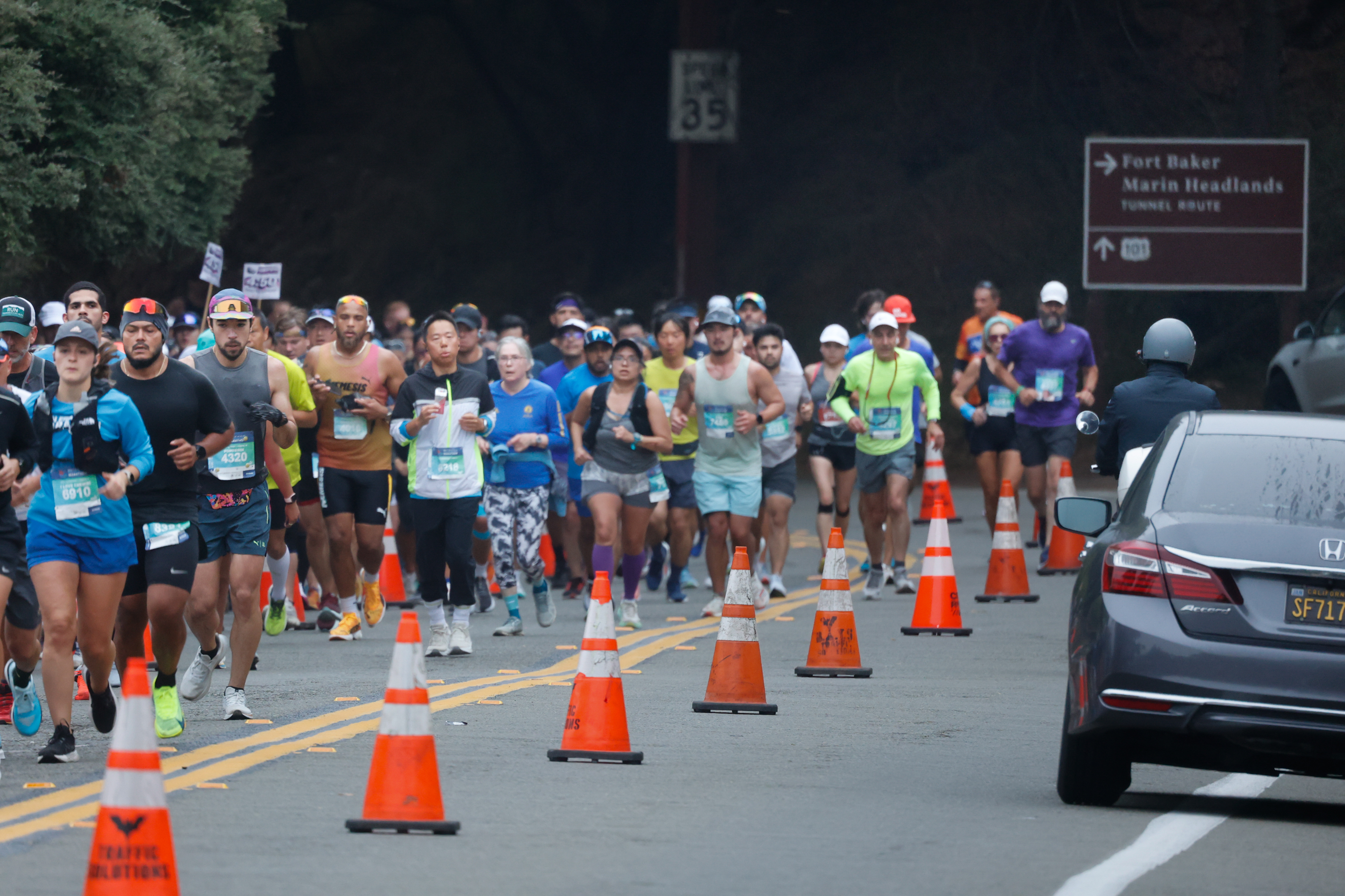 A group of runners in athletic gear is participating in a race on a road with traffic cones, while a motorbike rider and car are visible to the right.