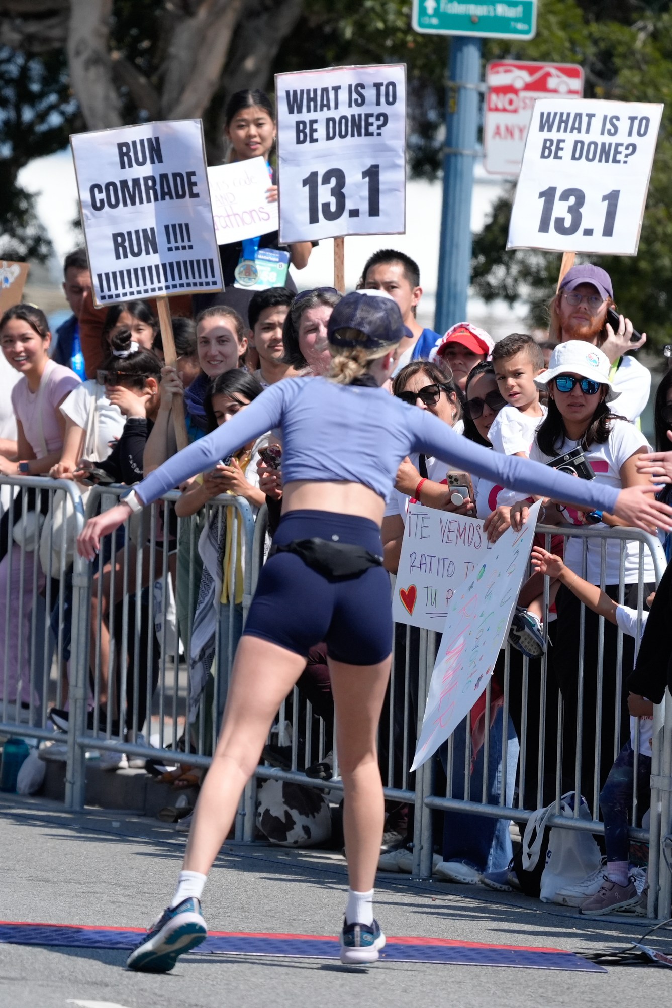 A runner nears the finish line, reaching out to spectators behind barriers holding supportive signs, including “Run Comrade Run” and “What is to be done? 13.1.”