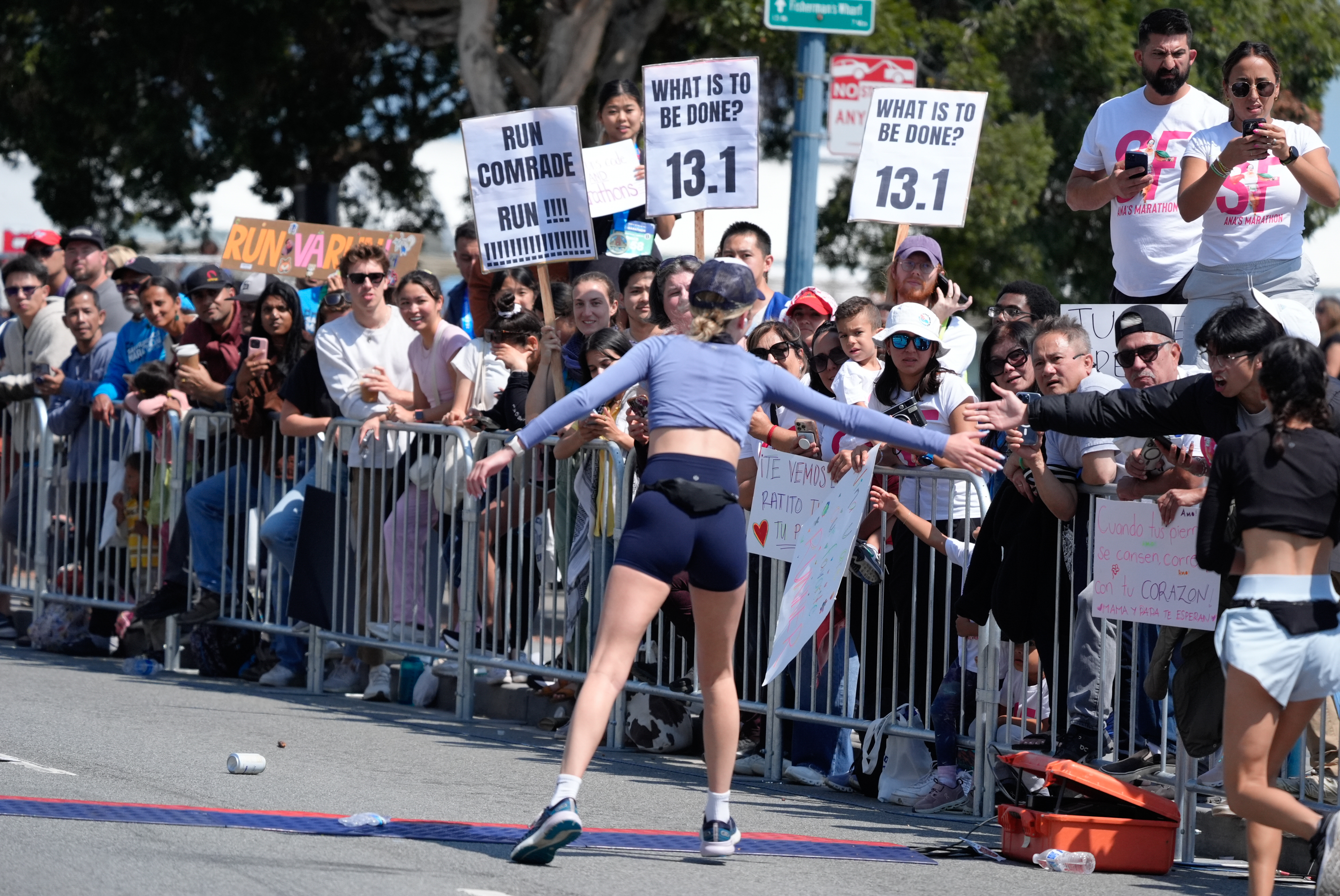 A runner nears the finish line, reaching out to spectators behind barriers holding supportive signs, including “Run Comrade Run” and “What is to be done? 13.1.”