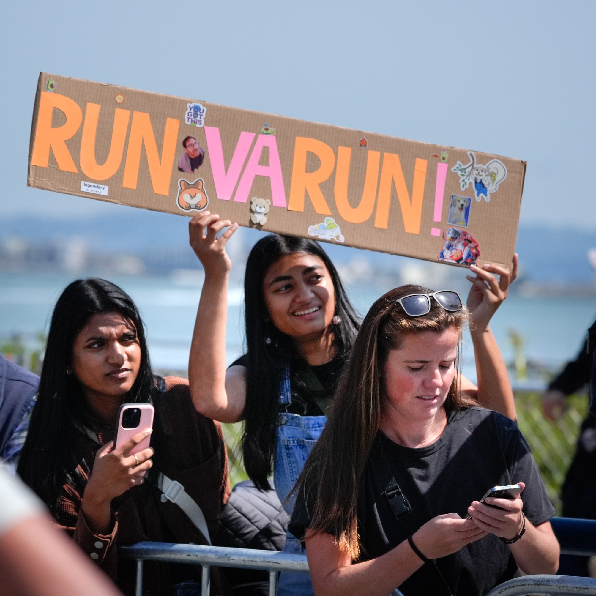 A group of people are watching an event outdoors, with one person holding a sign that says &quot;RUN VARUN RUN!&quot; while another looks at their phone.