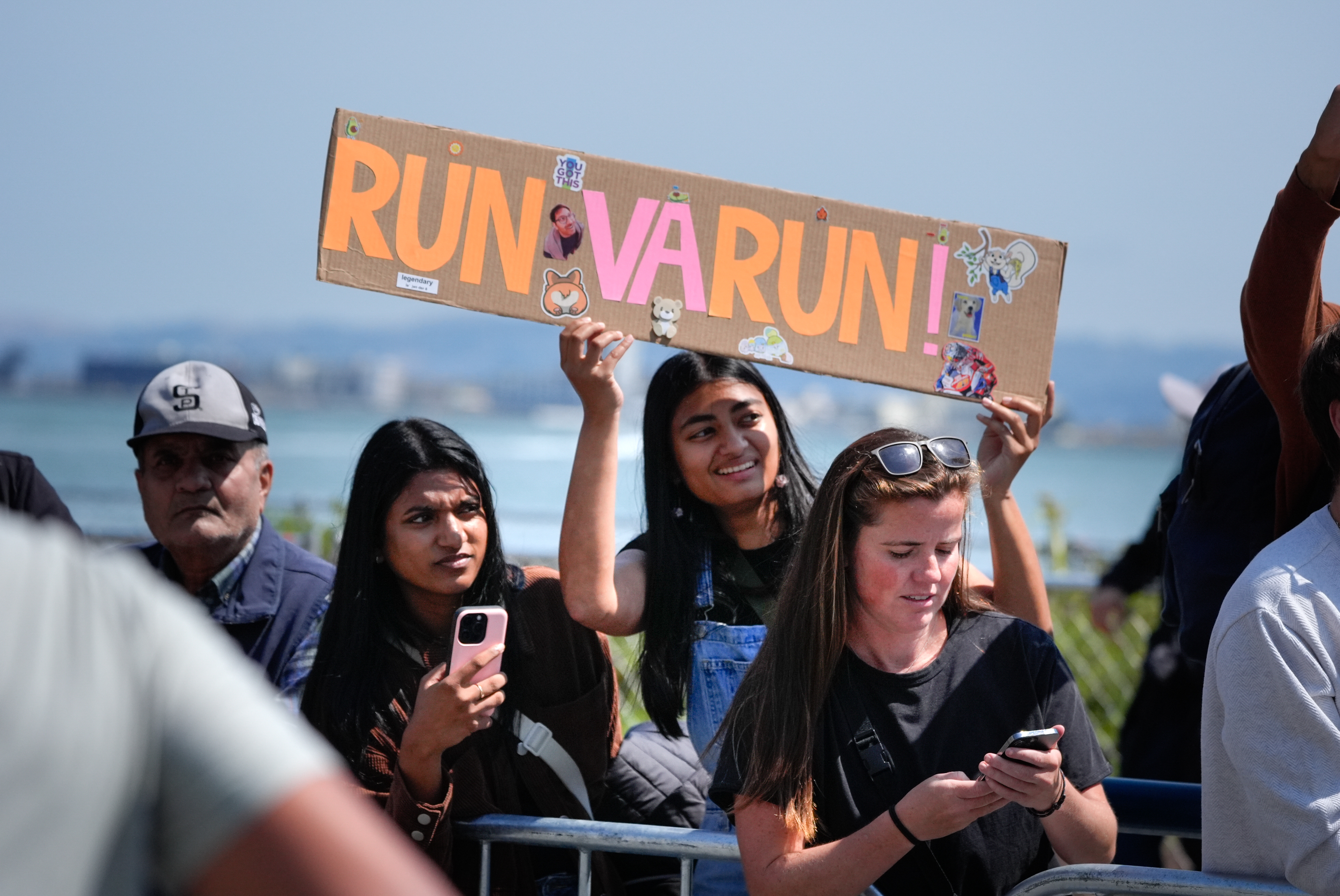 A group of people are watching an event outdoors, with one person holding a sign that says &quot;RUN VARUN RUN!&quot; while another looks at their phone.