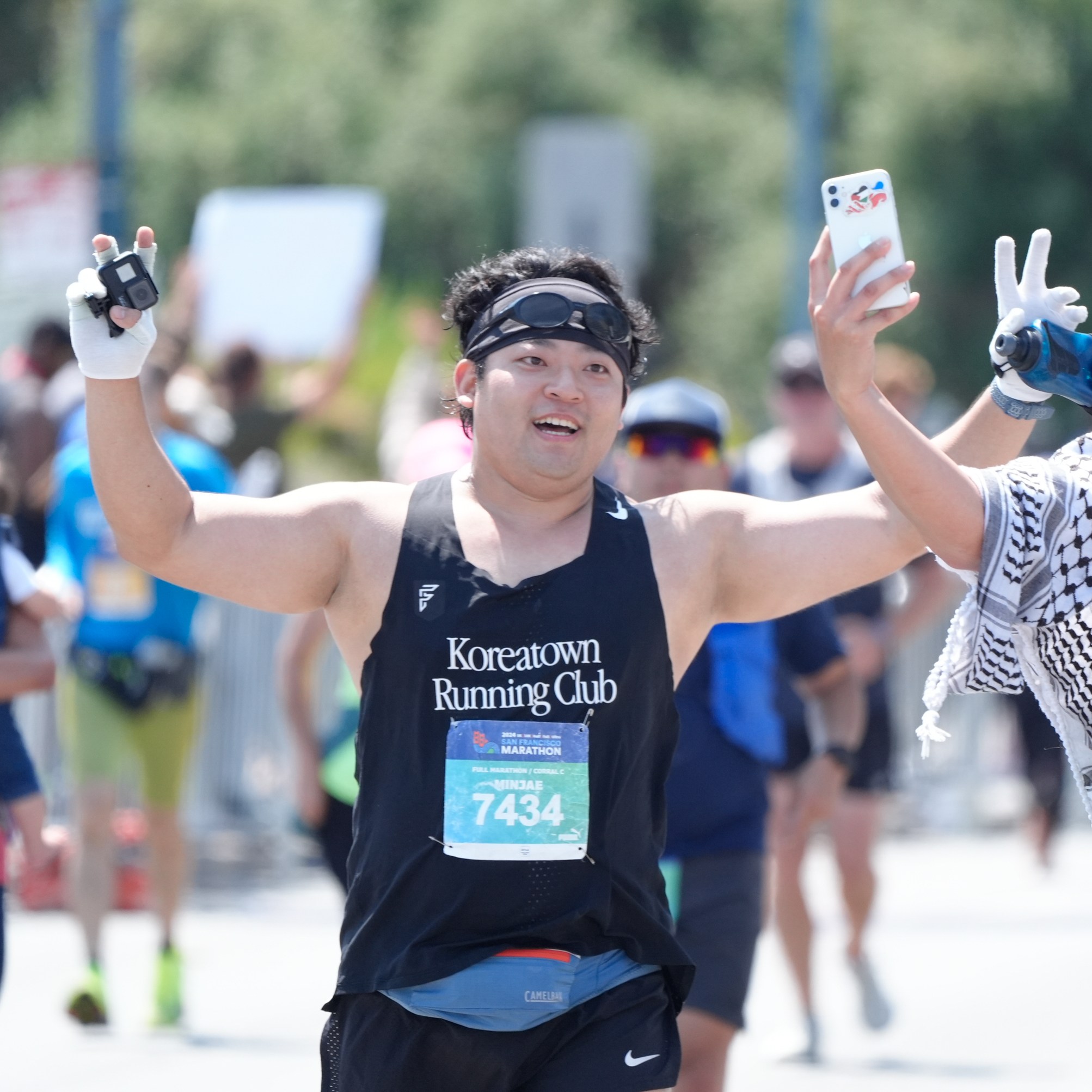 Two marathon runners, smiling and raising their hands, are taking a selfie. One wears a &quot;Koreatown Running Club&quot; shirt and the other has a &quot;K/R/C&quot; shirt and a striped scarf.