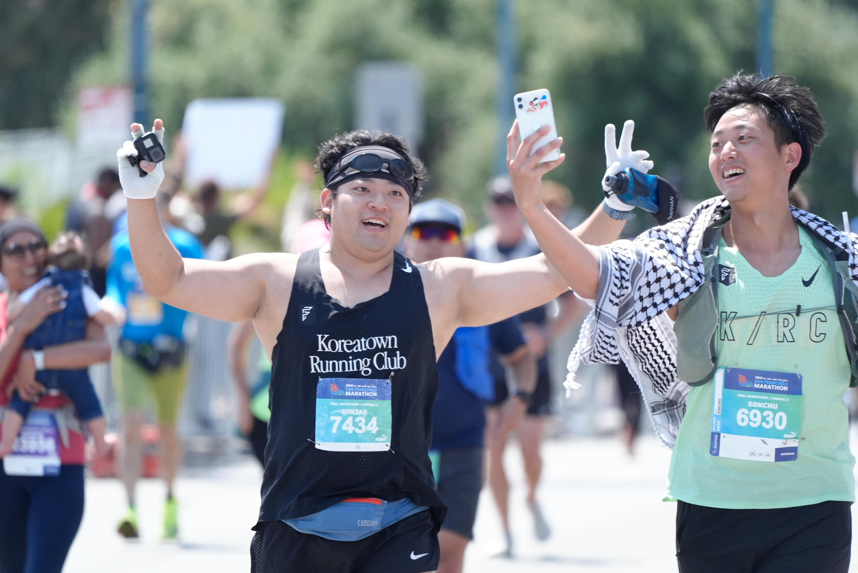 Two marathon runners, smiling and raising their hands, are taking a selfie. One wears a &quot;Koreatown Running Club&quot; shirt and the other has a &quot;K/R/C&quot; shirt and a striped scarf.