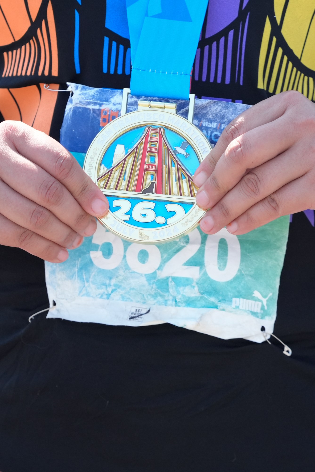 A person wearing a black shirt and a fitness tracker holds a marathon medal labeled &quot;26.2&quot; with an image of the Golden Gate Bridge and a bib number &quot;5820&quot;.