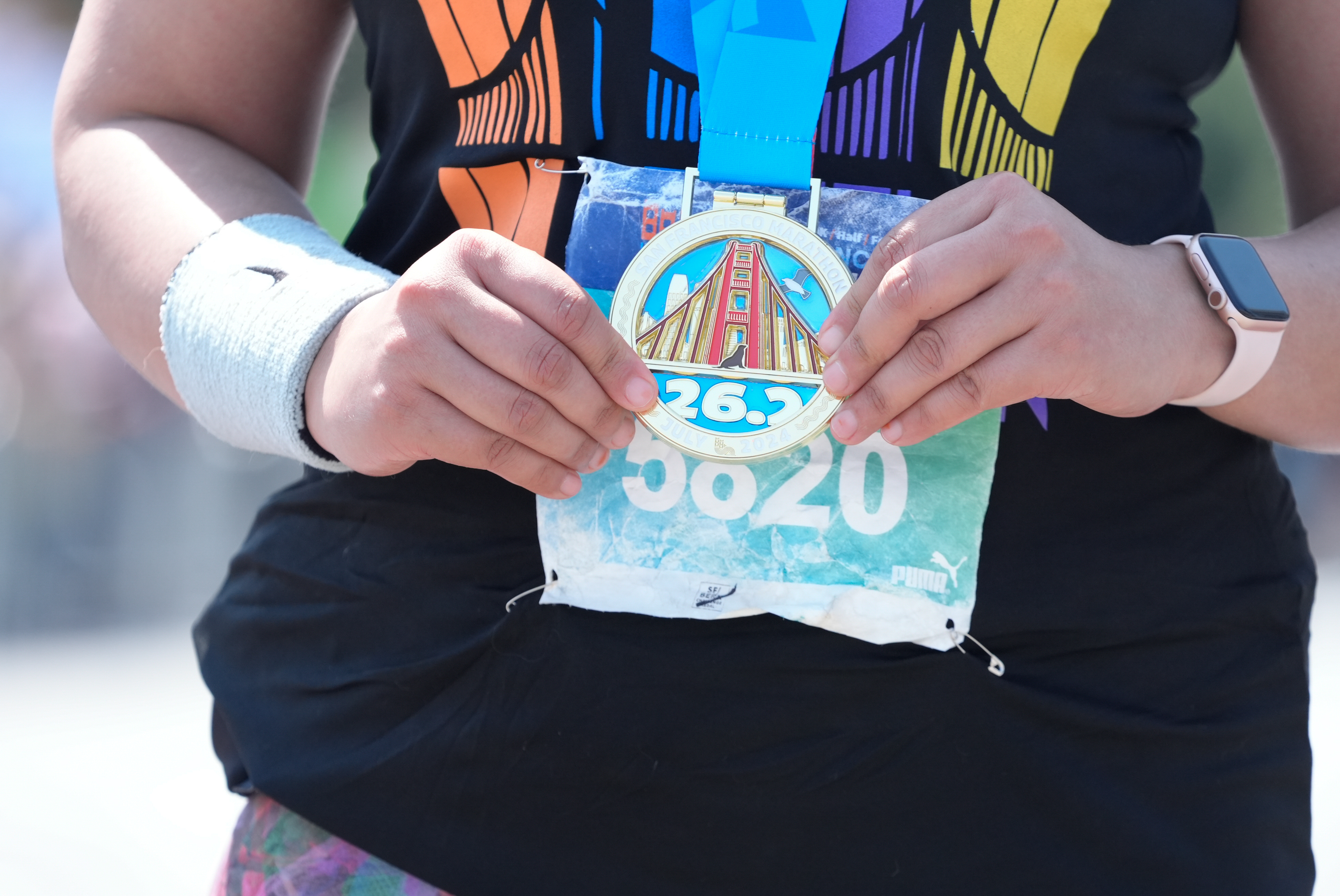 A person wearing a black shirt and a fitness tracker holds a marathon medal labeled &quot;26.2&quot; with an image of the Golden Gate Bridge and a bib number &quot;5820&quot;.