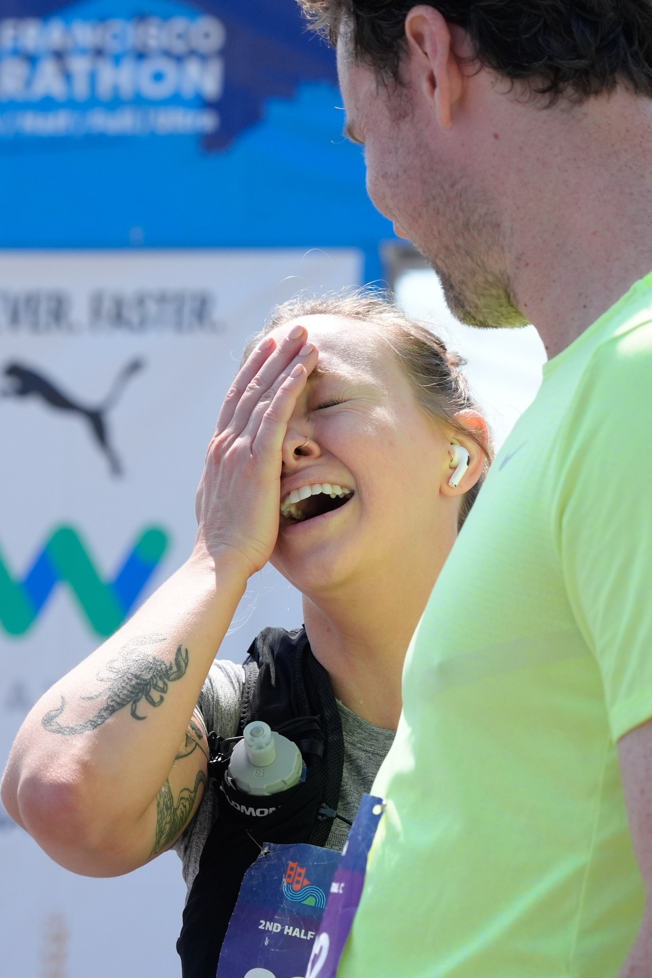 A woman laughs while covering her face, standing beside a man in a neon yellow shirt at the San Francisco Marathon finish line, with a blue marathon banner in the background.