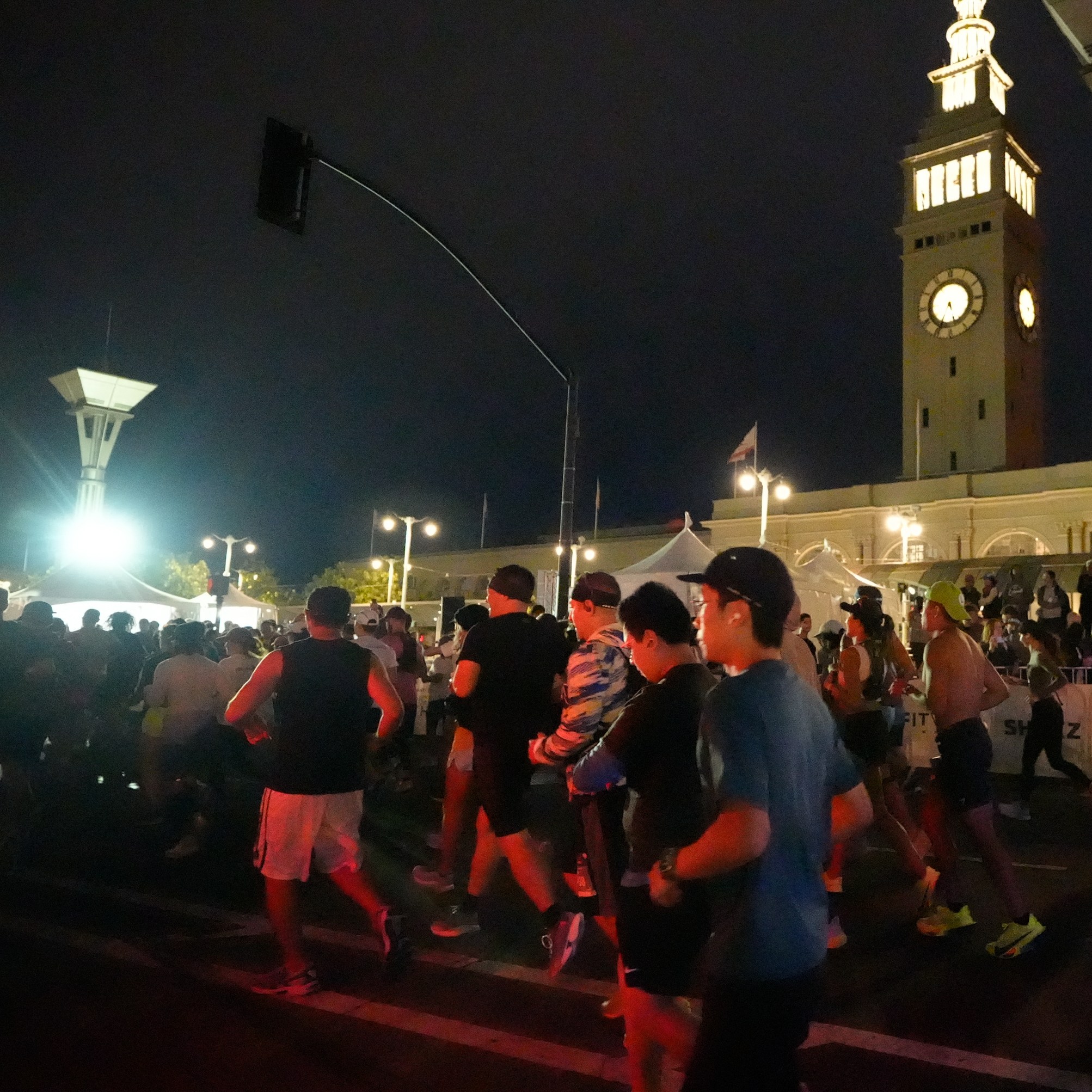 A group of runners is participating in a nighttime race, with an illuminated clock tower in the background. The scene is lively with people wearing athletic gear.