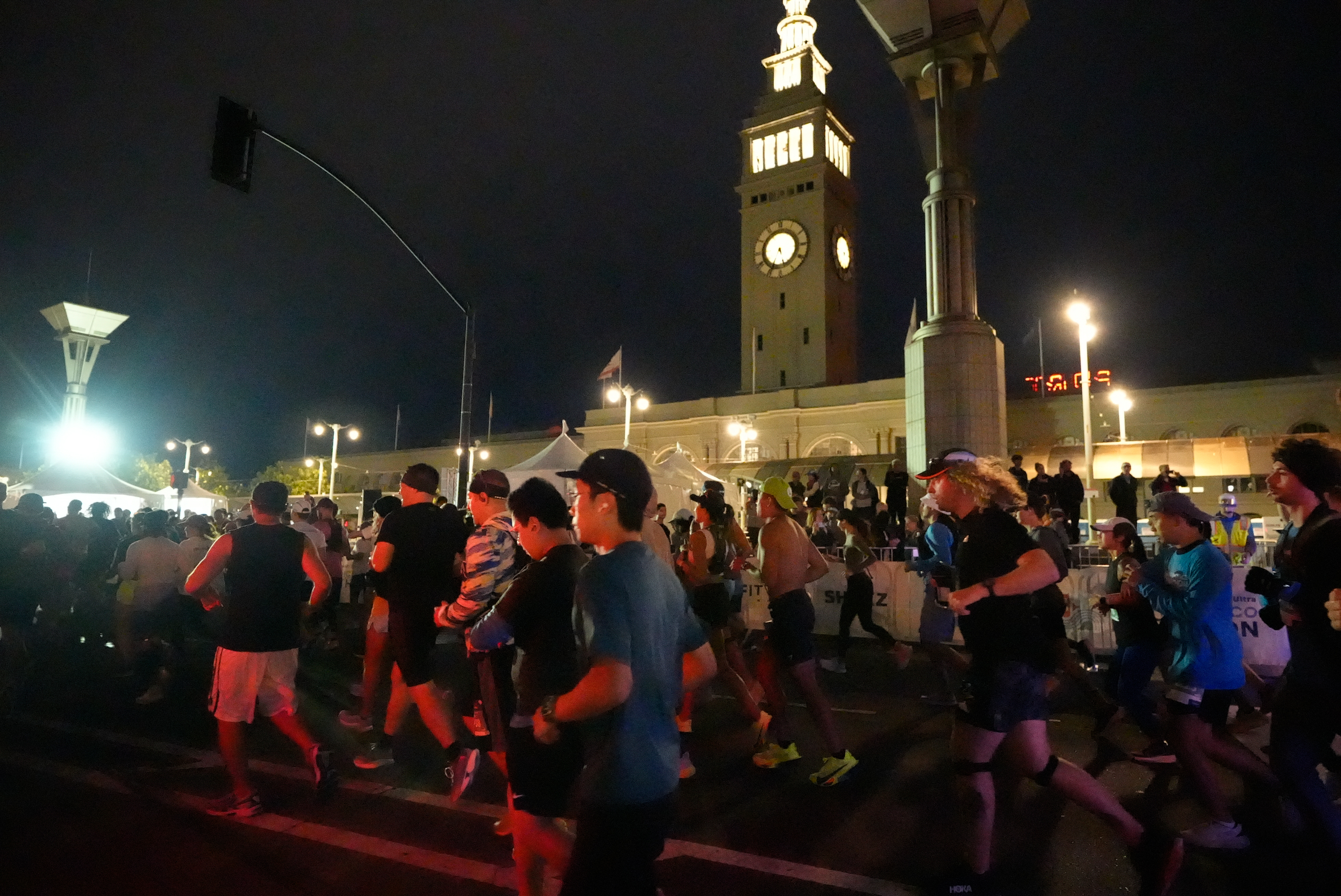 A group of runners is participating in a nighttime race, with an illuminated clock tower in the background. The scene is lively with people wearing athletic gear.