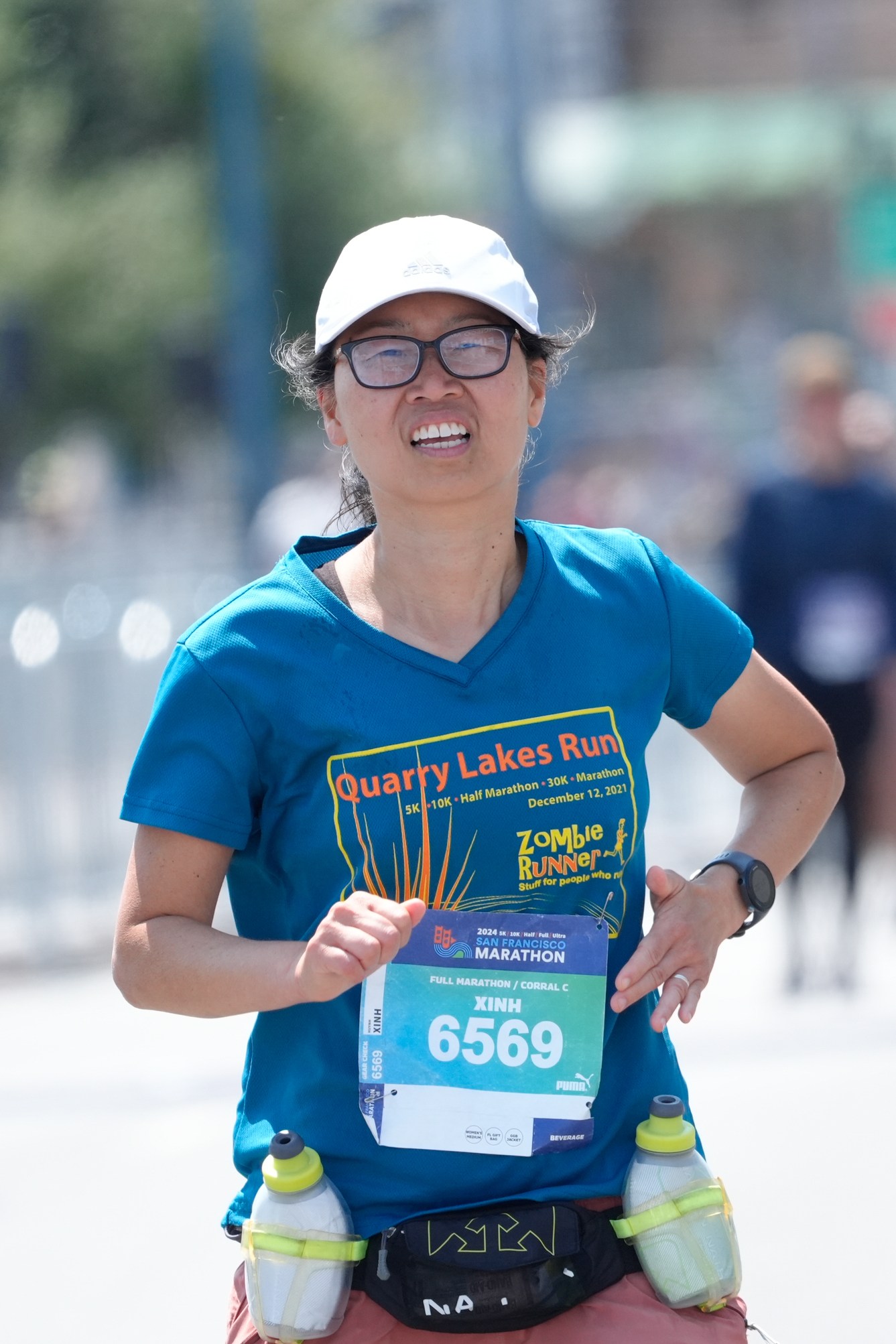 A runner in a blue &quot;Quarry Lakes Run&quot; shirt with race number 6569 is running outdoors, wearing a white cap and glasses, with two water bottles strapped to her waist.