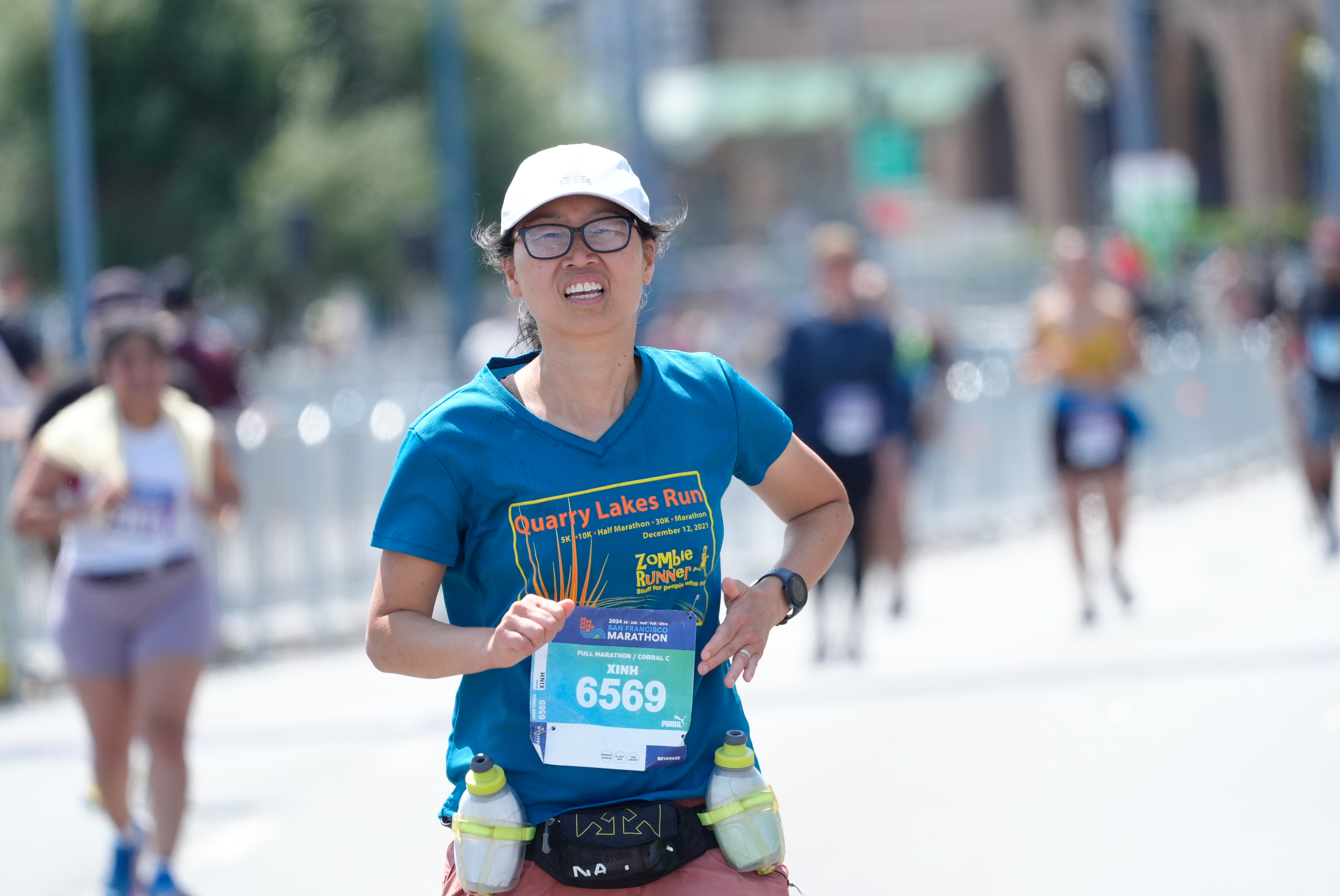 A runner in a blue &quot;Quarry Lakes Run&quot; shirt with race number 6569 is running outdoors, wearing a white cap and glasses, with two water bottles strapped to her waist.