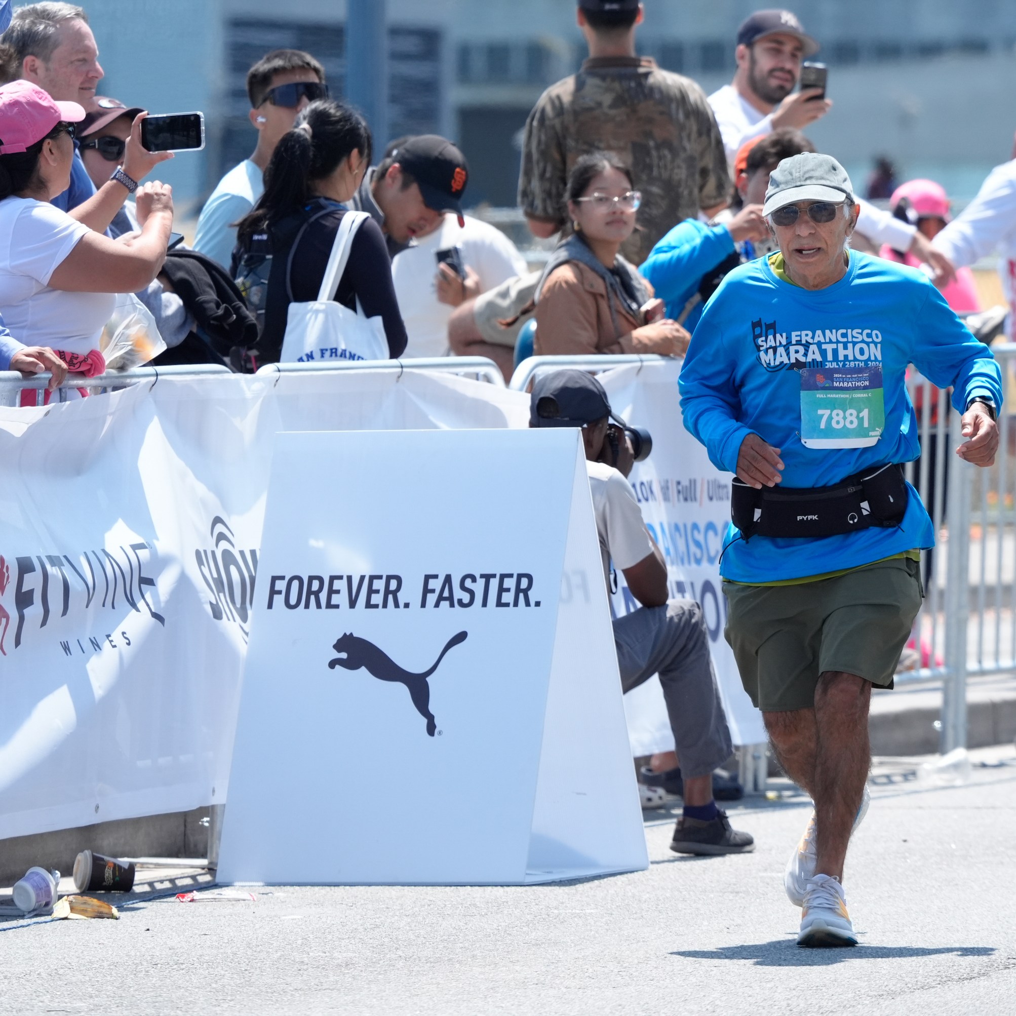 A marathon runner in a blue shirt and khaki shorts nears the finish line, cheered by a crowd behind metal barriers. A sign reads &quot;FOREVER. FASTER.&quot; with the Puma logo.