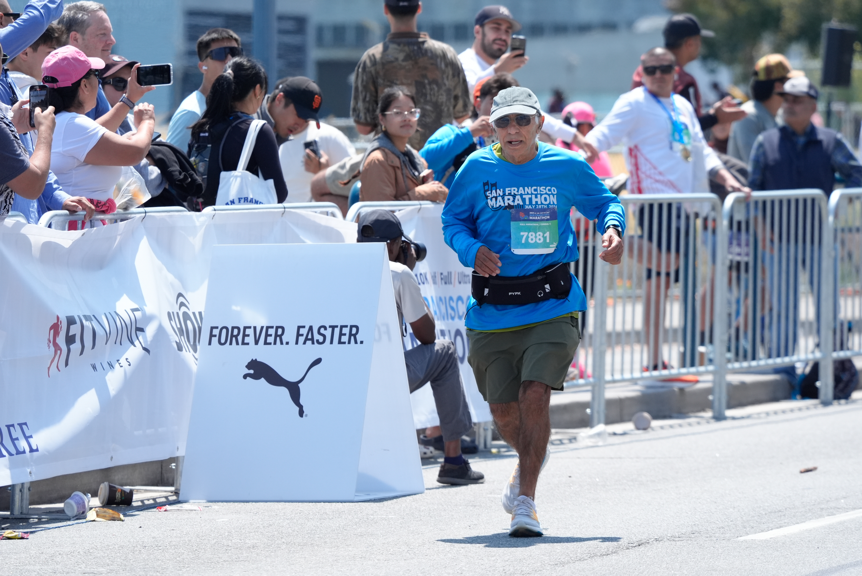 A marathon runner in a blue shirt and khaki shorts nears the finish line, cheered by a crowd behind metal barriers. A sign reads &quot;FOREVER. FASTER.&quot; with the Puma logo.