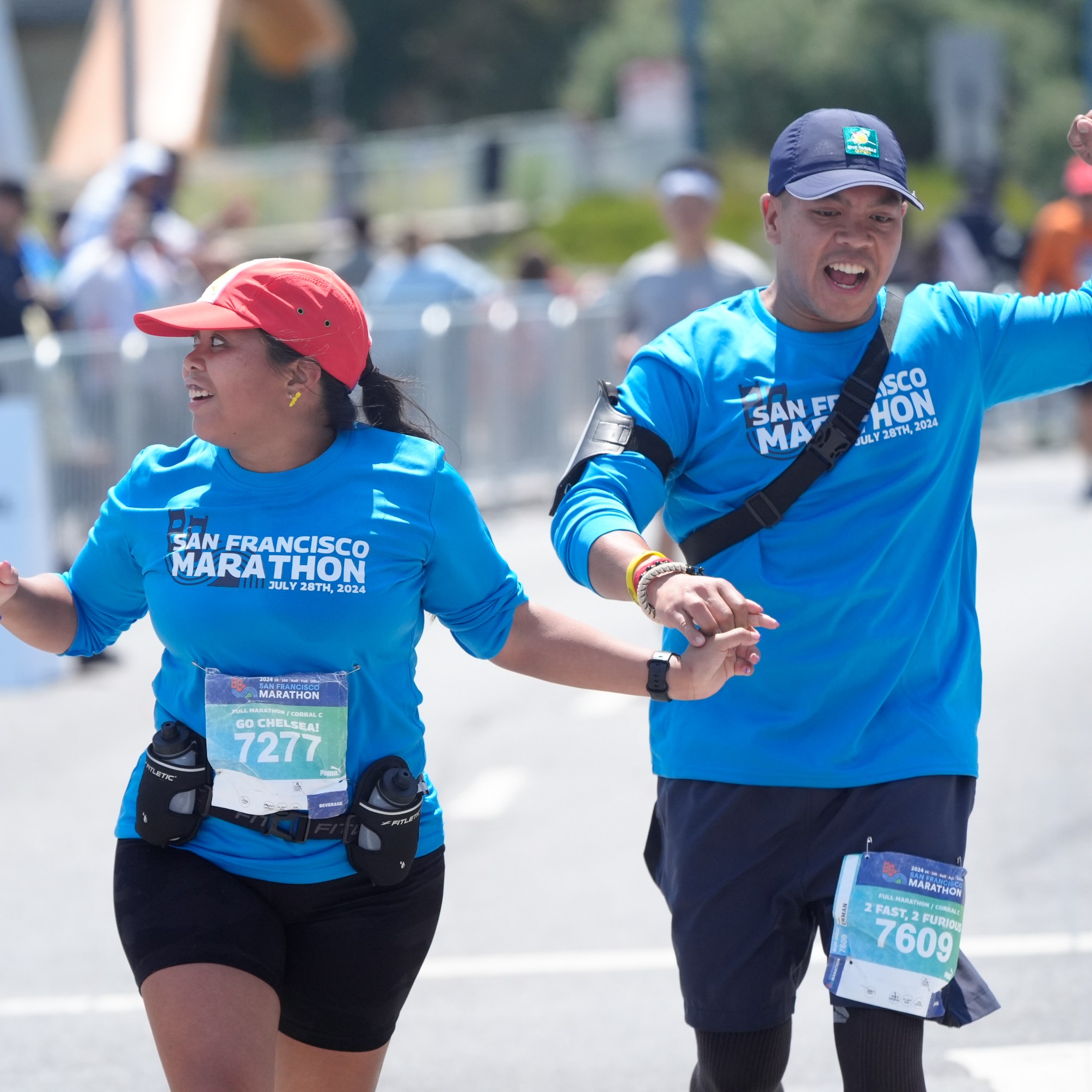 Two marathon runners, wearing blue &quot;San Francisco Marathon&quot; shirts, joyfully hold hands while running, surrounded by spectators and fellow participants.