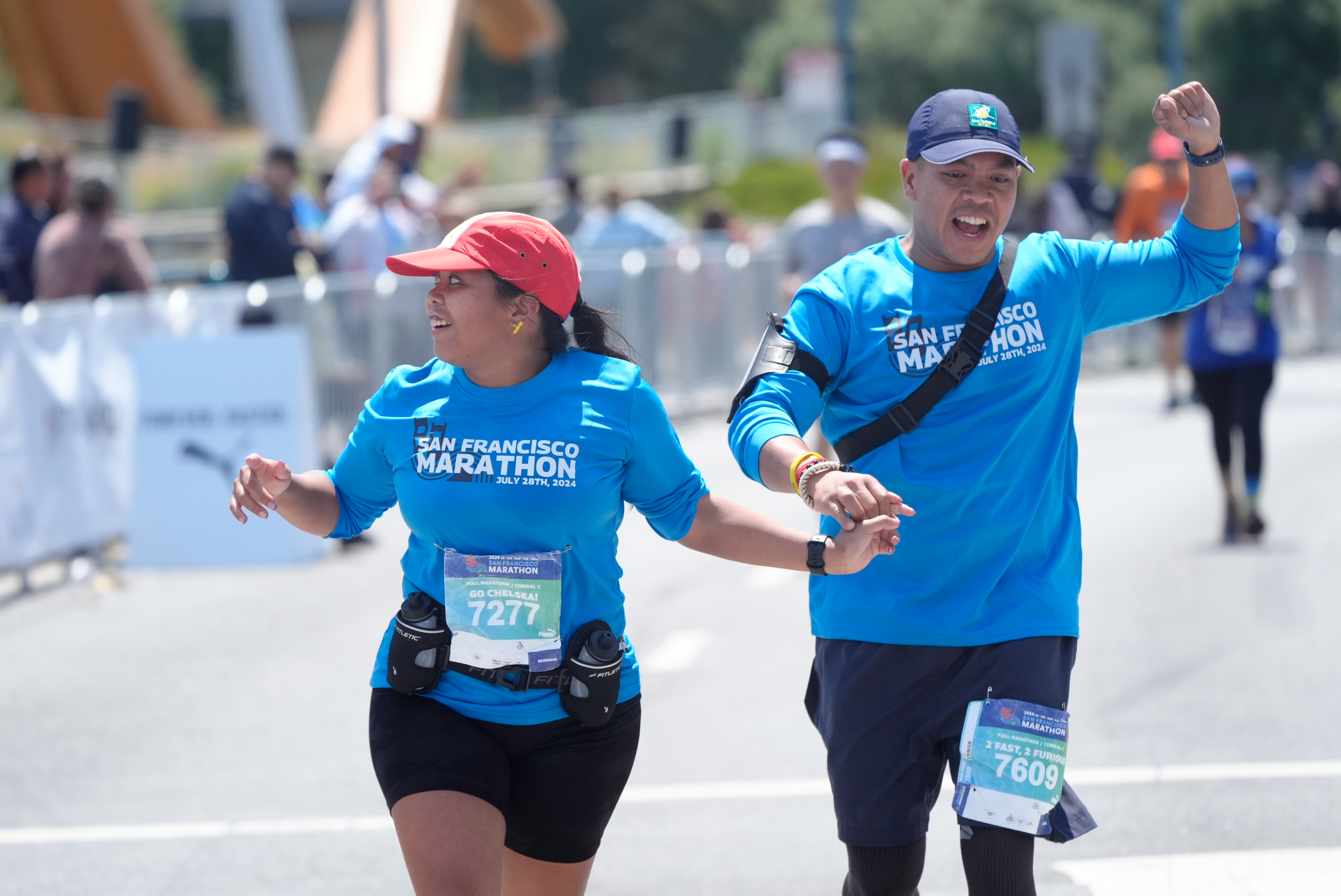 Two marathon runners, wearing blue &quot;San Francisco Marathon&quot; shirts, joyfully hold hands while running, surrounded by spectators and fellow participants.