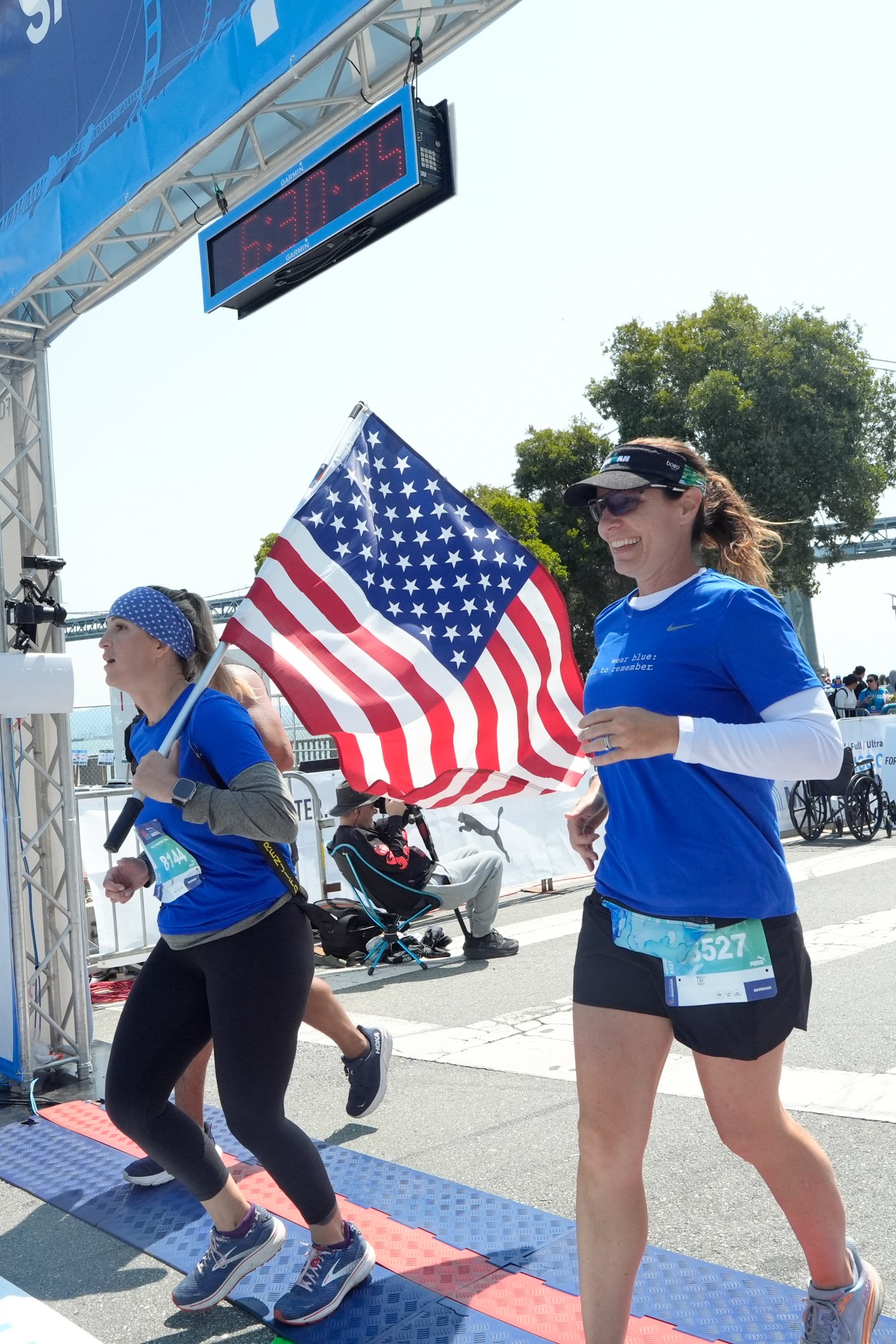 Two women in blue shirts, one holding an American flag, are crossing the finish line of a marathon, with a bridge and spectators in the background.