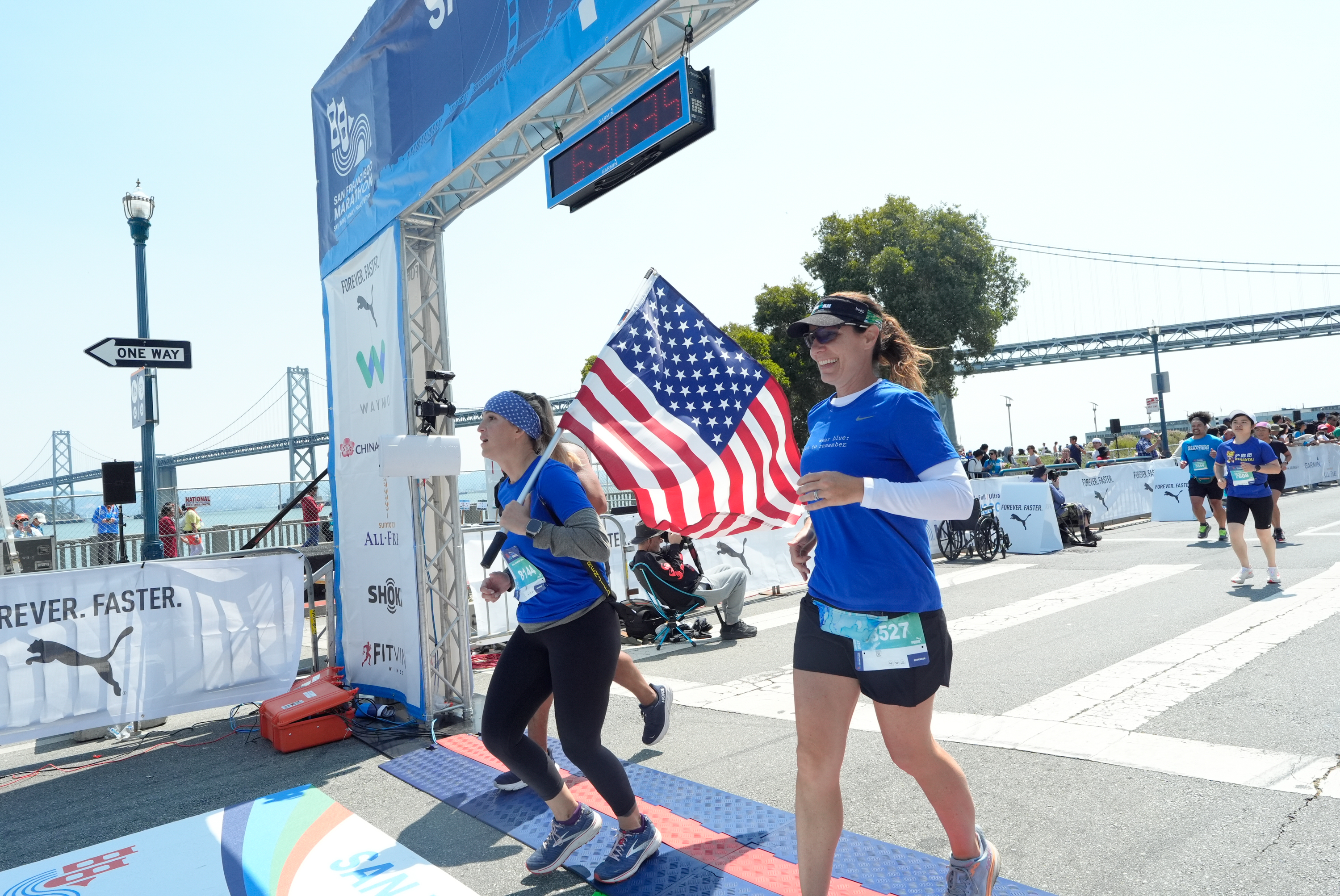 Two women in blue shirts, one holding an American flag, are crossing the finish line of a marathon, with a bridge and spectators in the background.