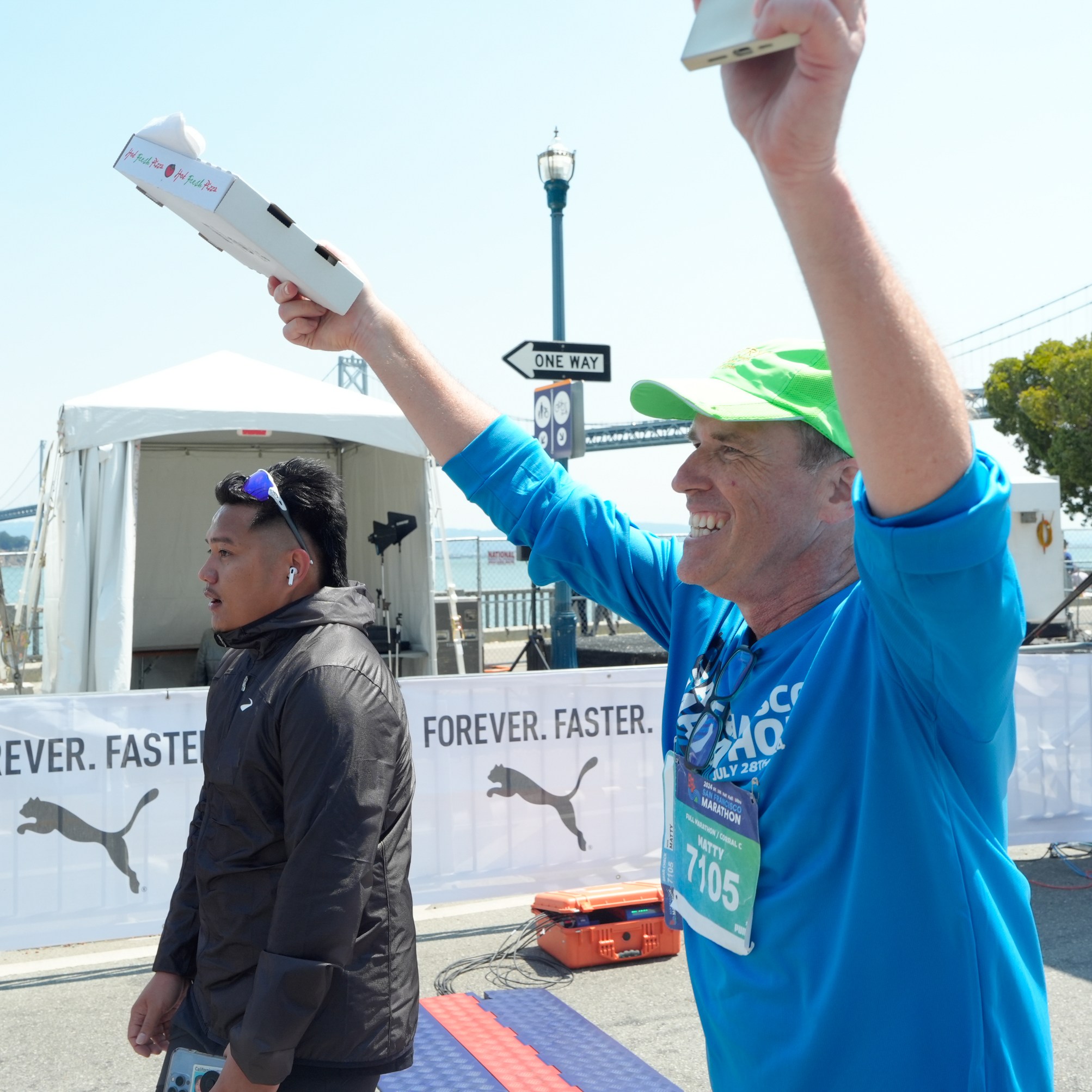 A man in a green cap and blue shirt celebrates at a marathon finish line, holding pizza boxes, while another man in a black jacket walks beside him.