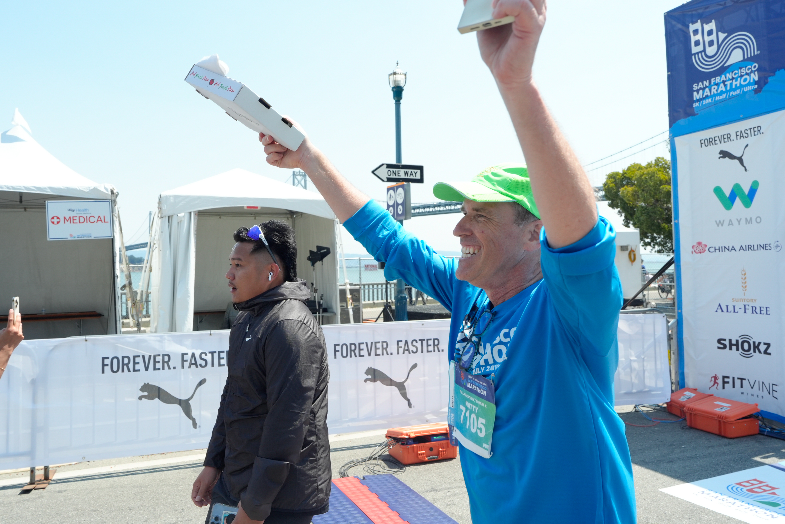 A man in a green cap and blue shirt celebrates at a marathon finish line, holding pizza boxes, while another man in a black jacket walks beside him.