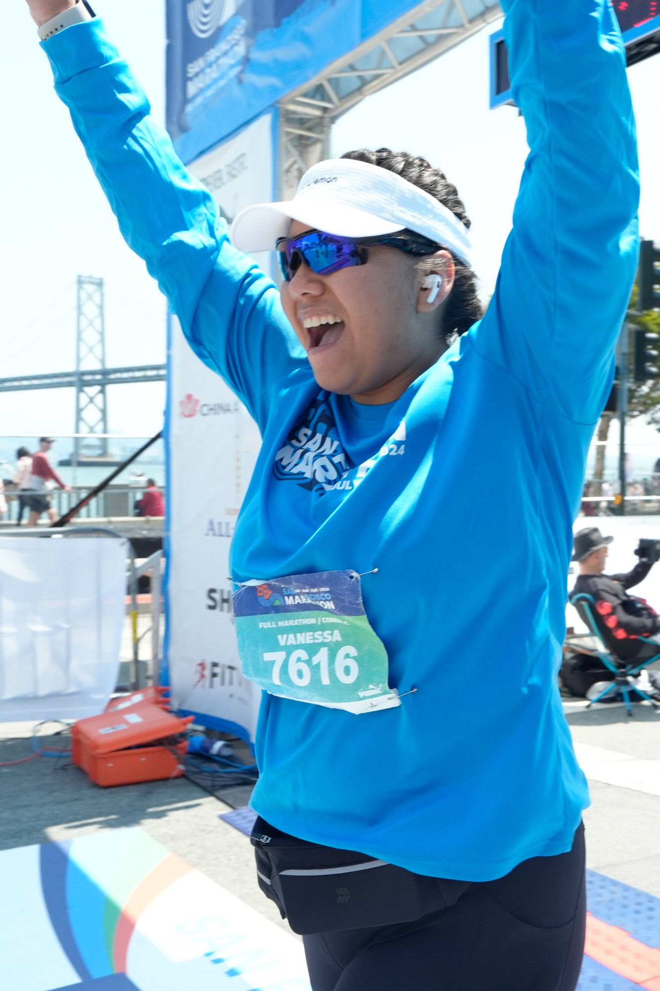 A joyful runner in a blue shirt, bib number 7616, and sunglasses, raises her arms in triumph at a marathon finish line, with a bridge and cheering spectators in the background.