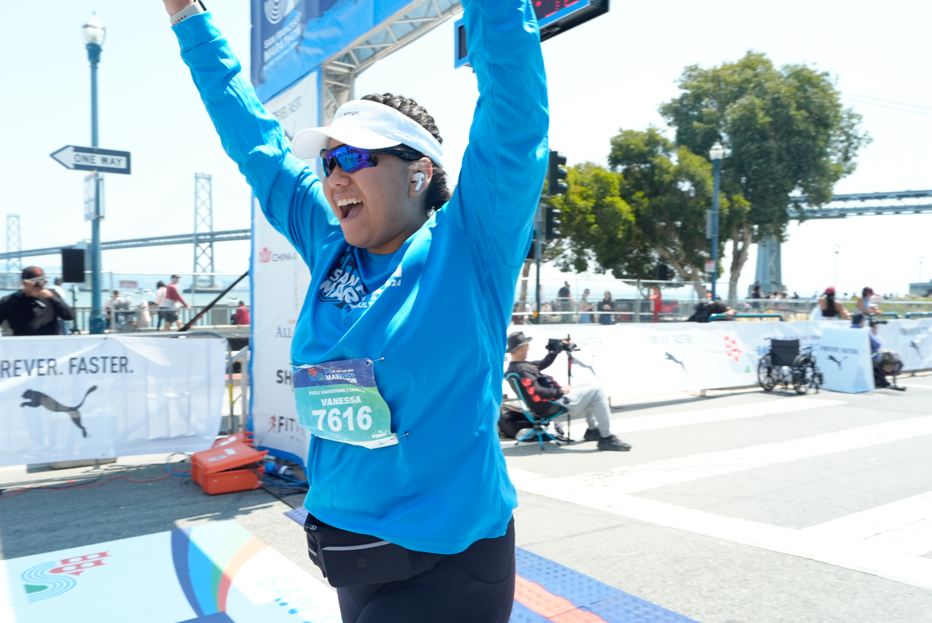 A joyful runner in a blue shirt, bib number 7616, and sunglasses, raises her arms in triumph at a marathon finish line, with a bridge and cheering spectators in the background.