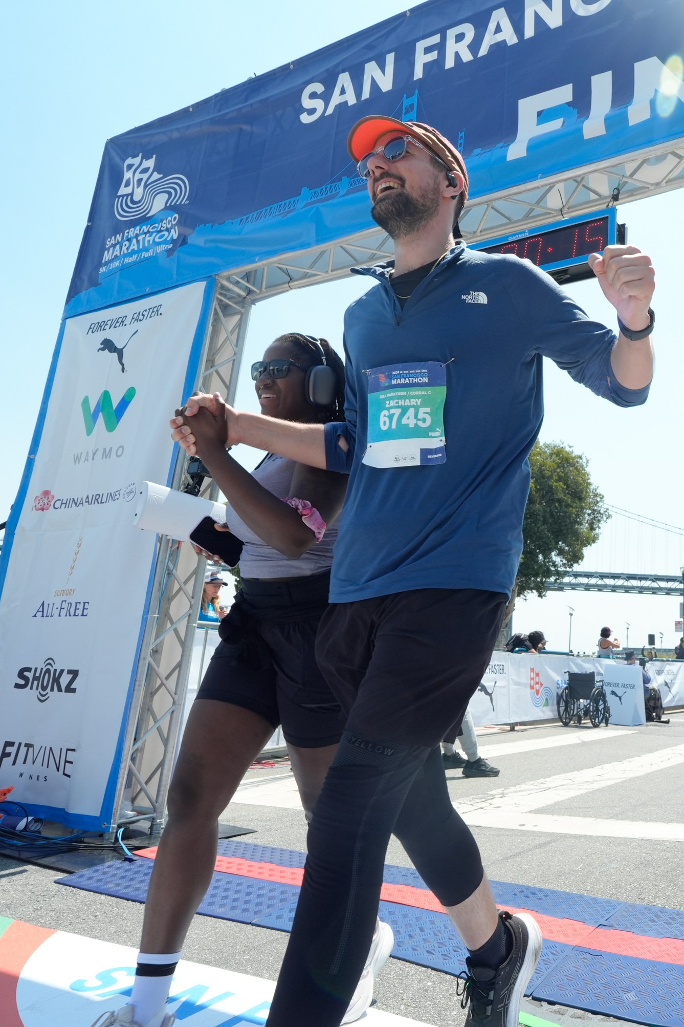 Two runners holding hands cross the finish line of the San Francisco Marathon, smiling. The race clock reads 3:45:29, and a bridge and spectators are visible in the background.